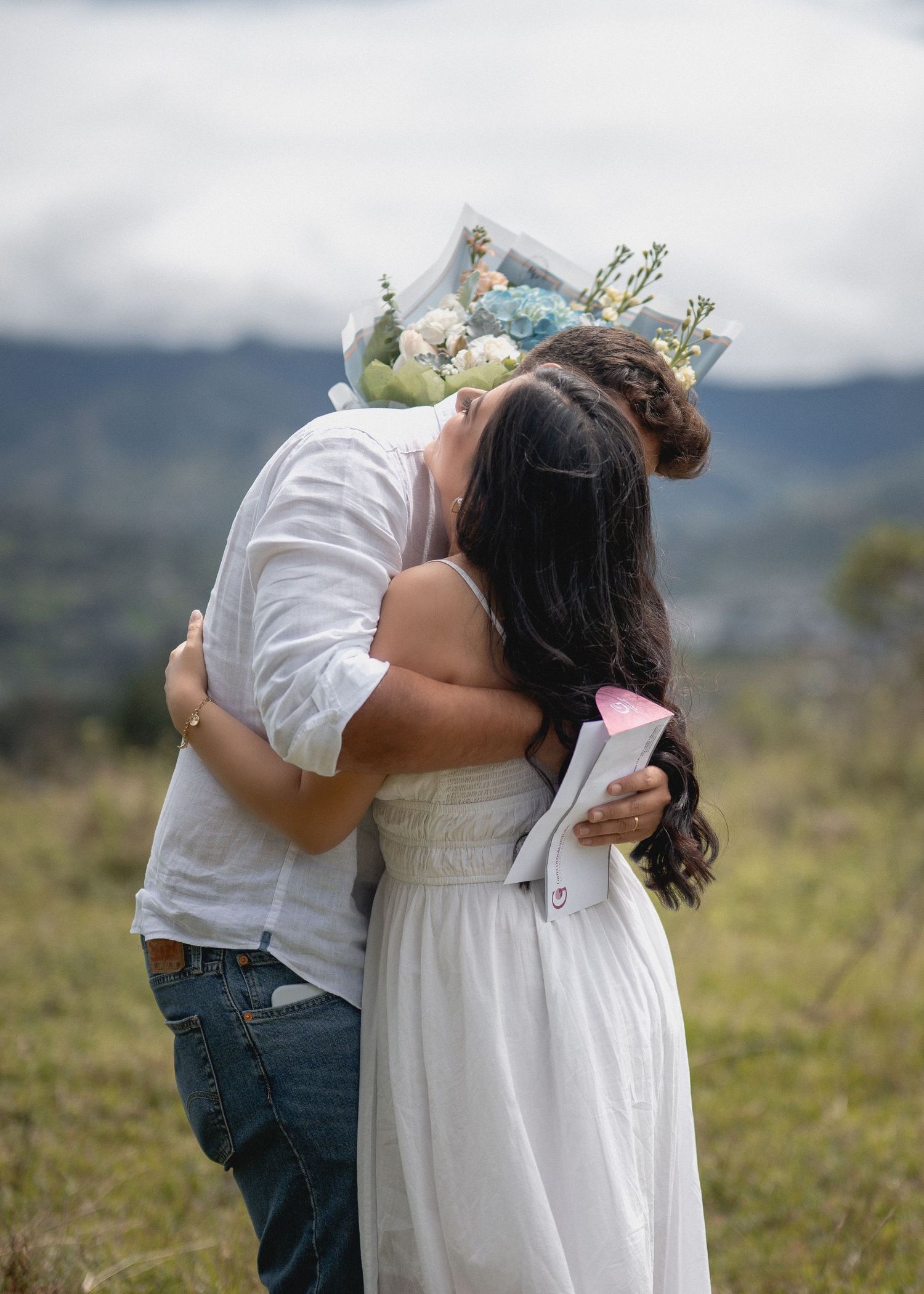 A couple embraces in a scenic outdoor setting with mountains and greenery in the background.