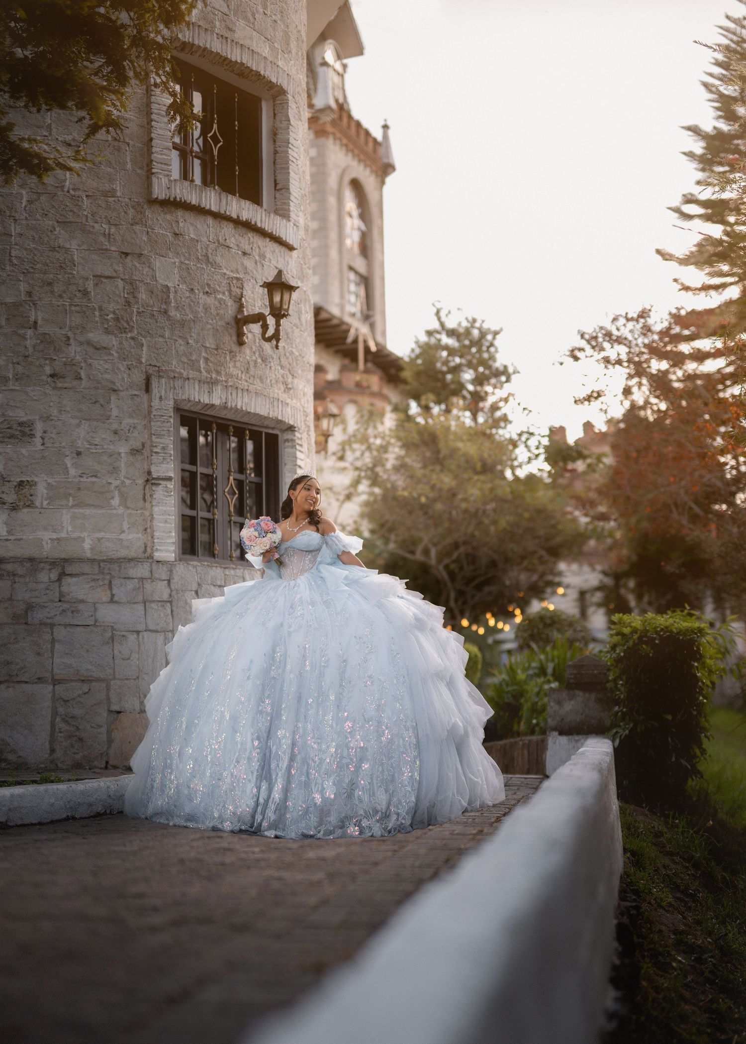 A bride in a voluminous white ball gown poses against a stone castle wall during sunset.