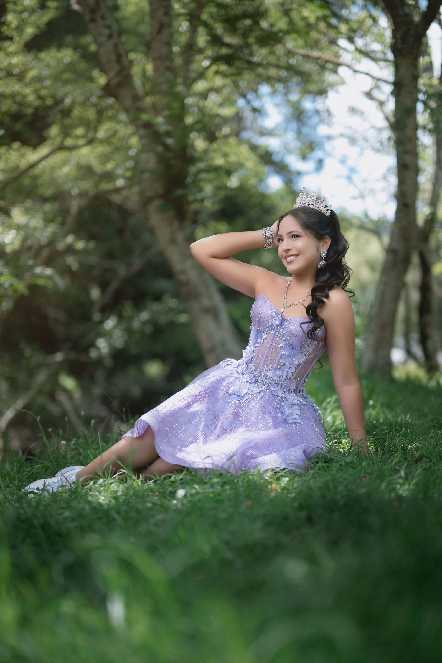 A person in a lavender formal dress reclines on green grass in a wooded outdoor setting.