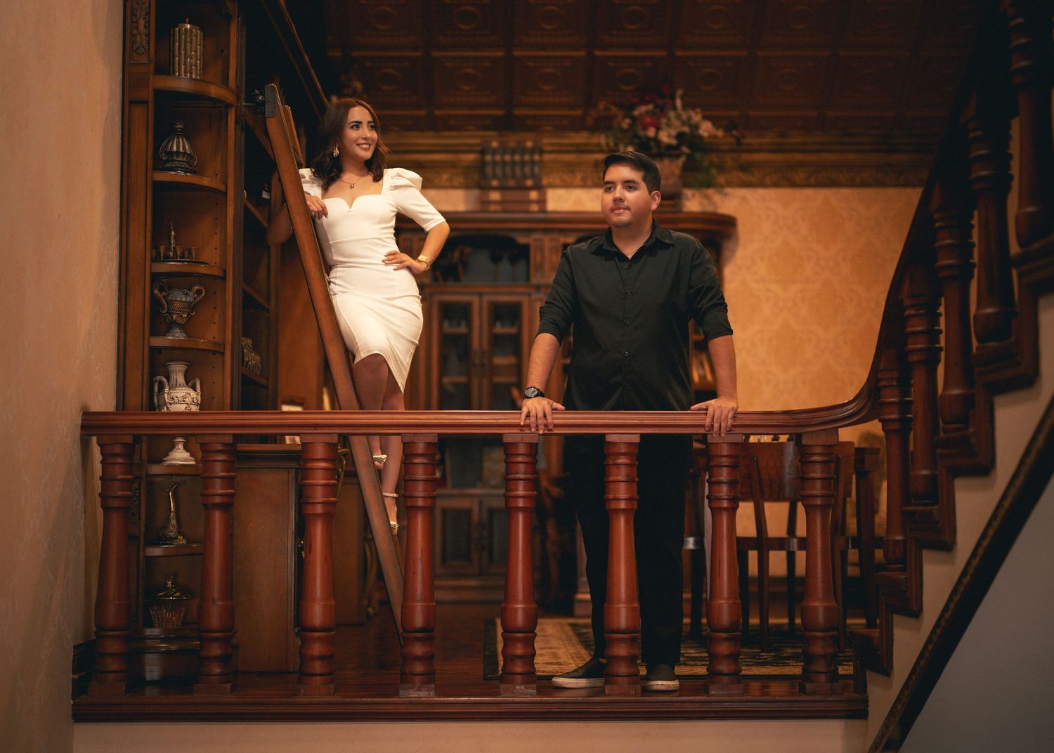 Couple poses on elegant wooden staircase during formal wedding photoshoot in warm-lit interior setting.