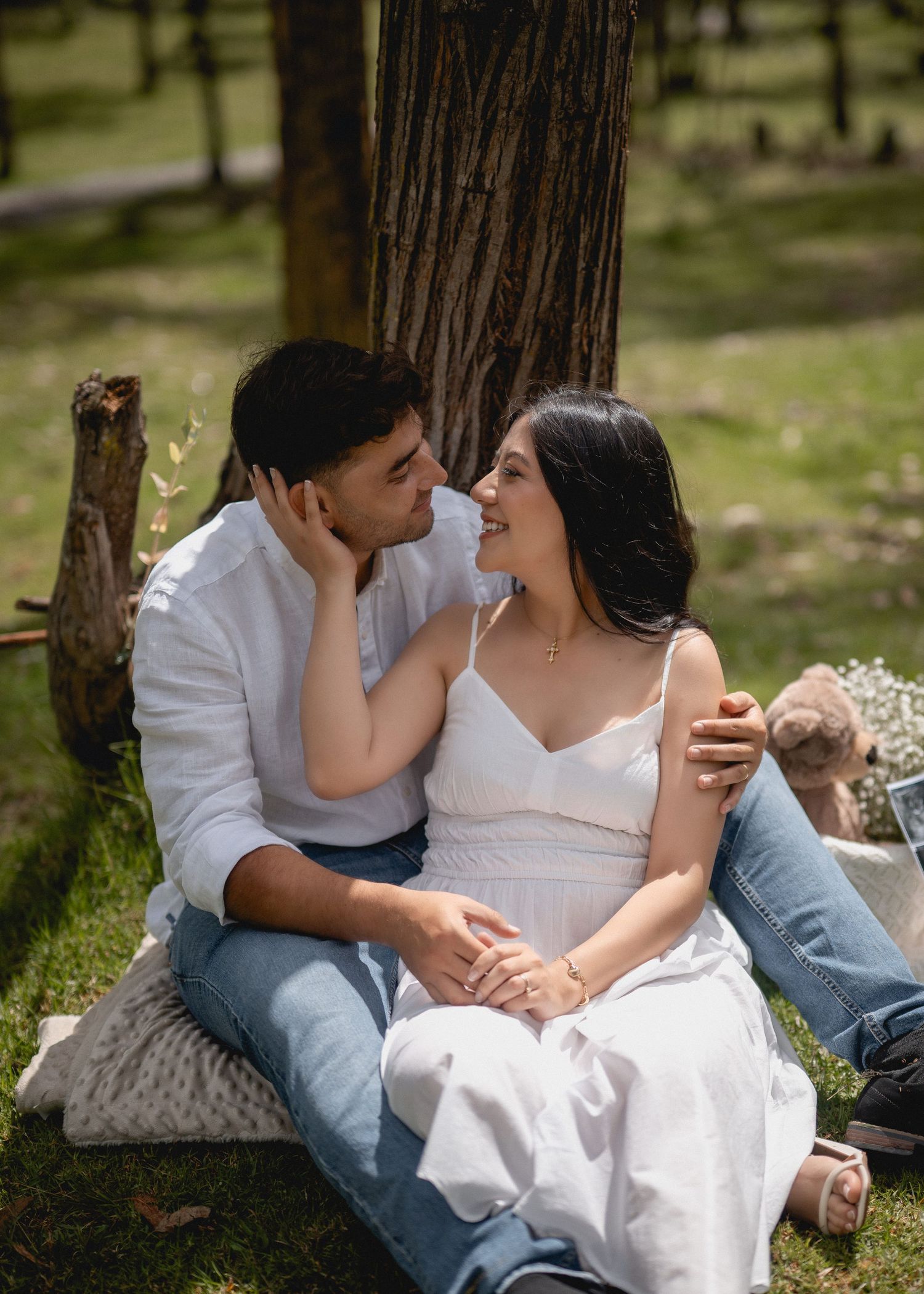 A couple in casual white and denim outfits share romantic moments while sitting together outdoors near trees.