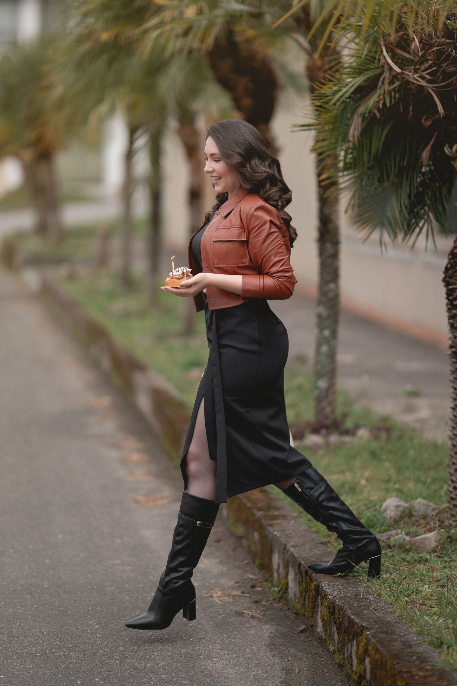 A woman in a brown leather jacket and black skirt walks along a palm tree-lined path wearing tall boots.