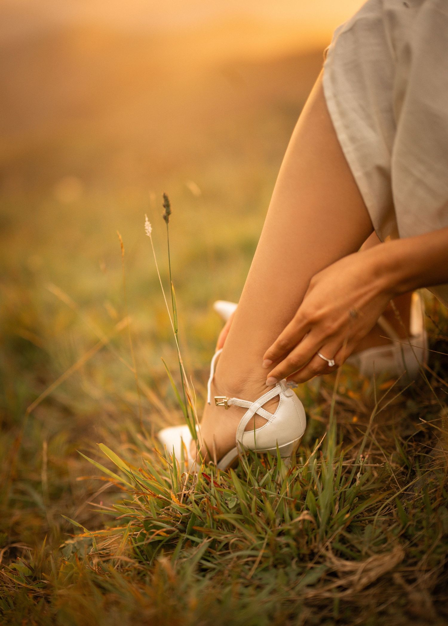 Close-up of white ballet flats on feet resting in sunlit grass during golden hour.