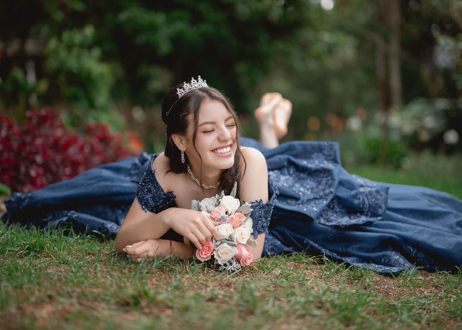 Someone in a navy blue formal gown lies on grass holding a pink and white bouquet while smiling outdoors.