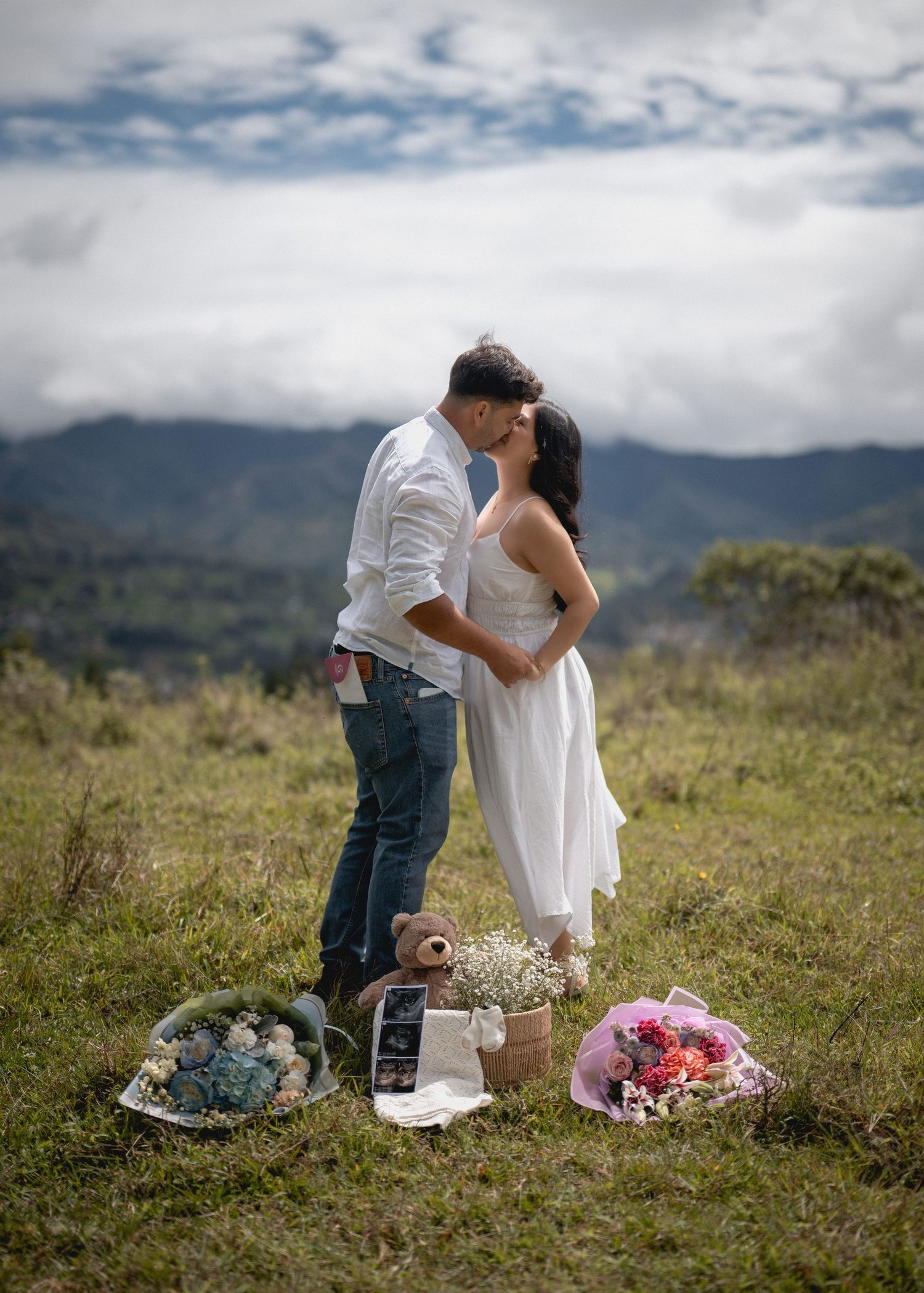 A romantic outdoor wedding proposal scene on a hilltop with flowers and decorations against a cloudy mountain backdrop.