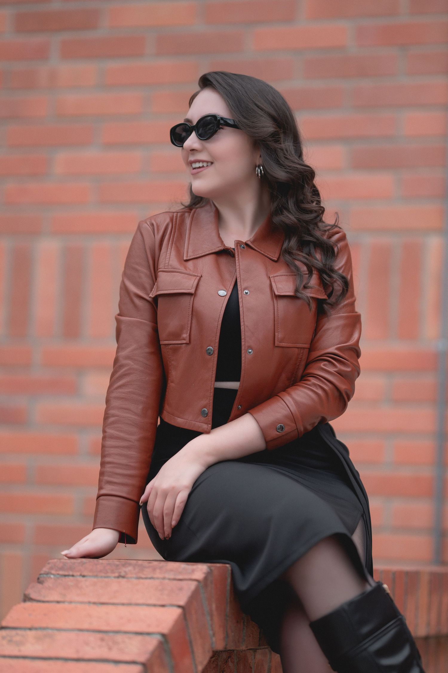 Model in brown leather jacket and black dress poses against brick wall in outdoor photoshoot.