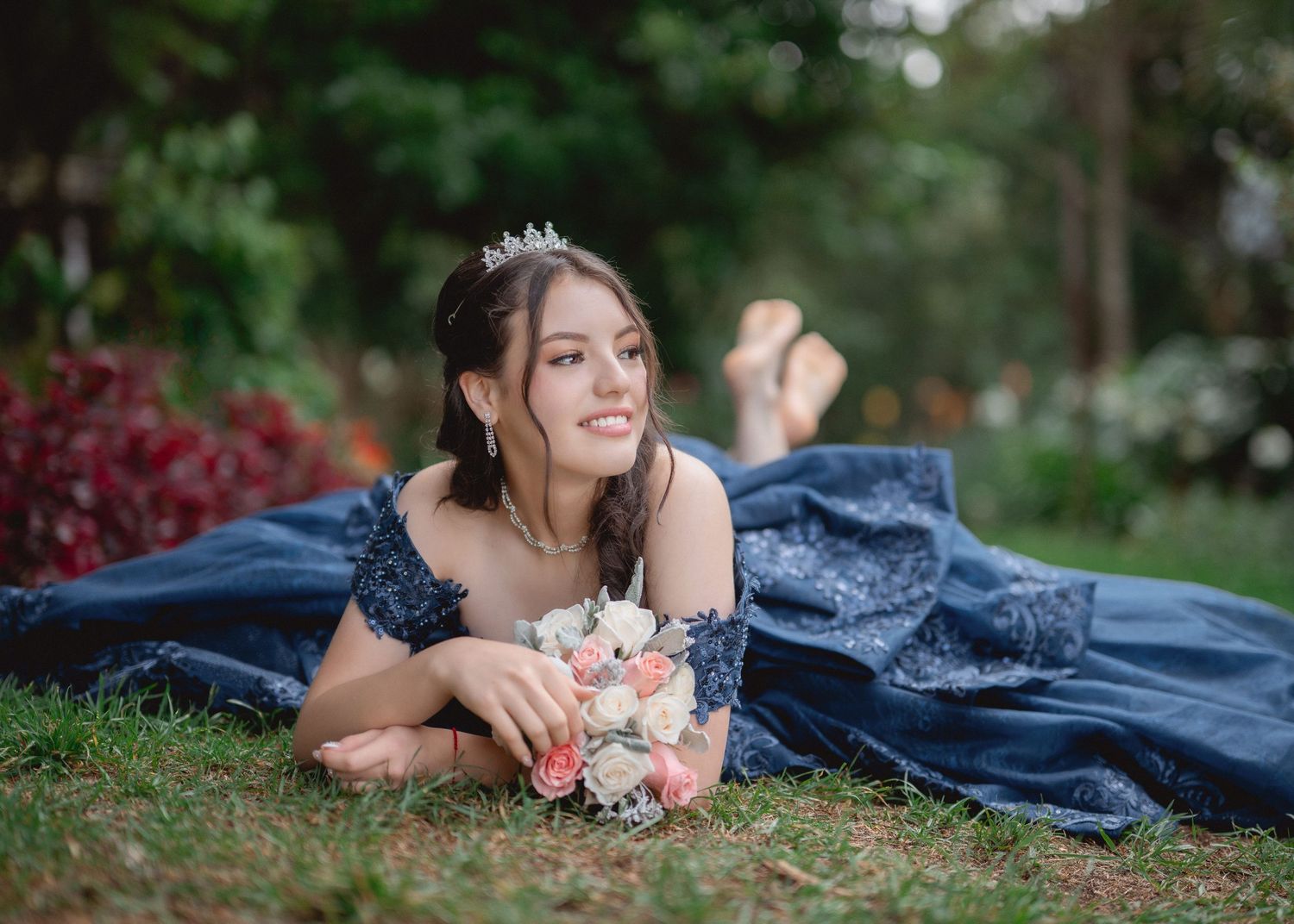 A woman in a blue dress lies on grass with pink flowers during an outdoor photo session.