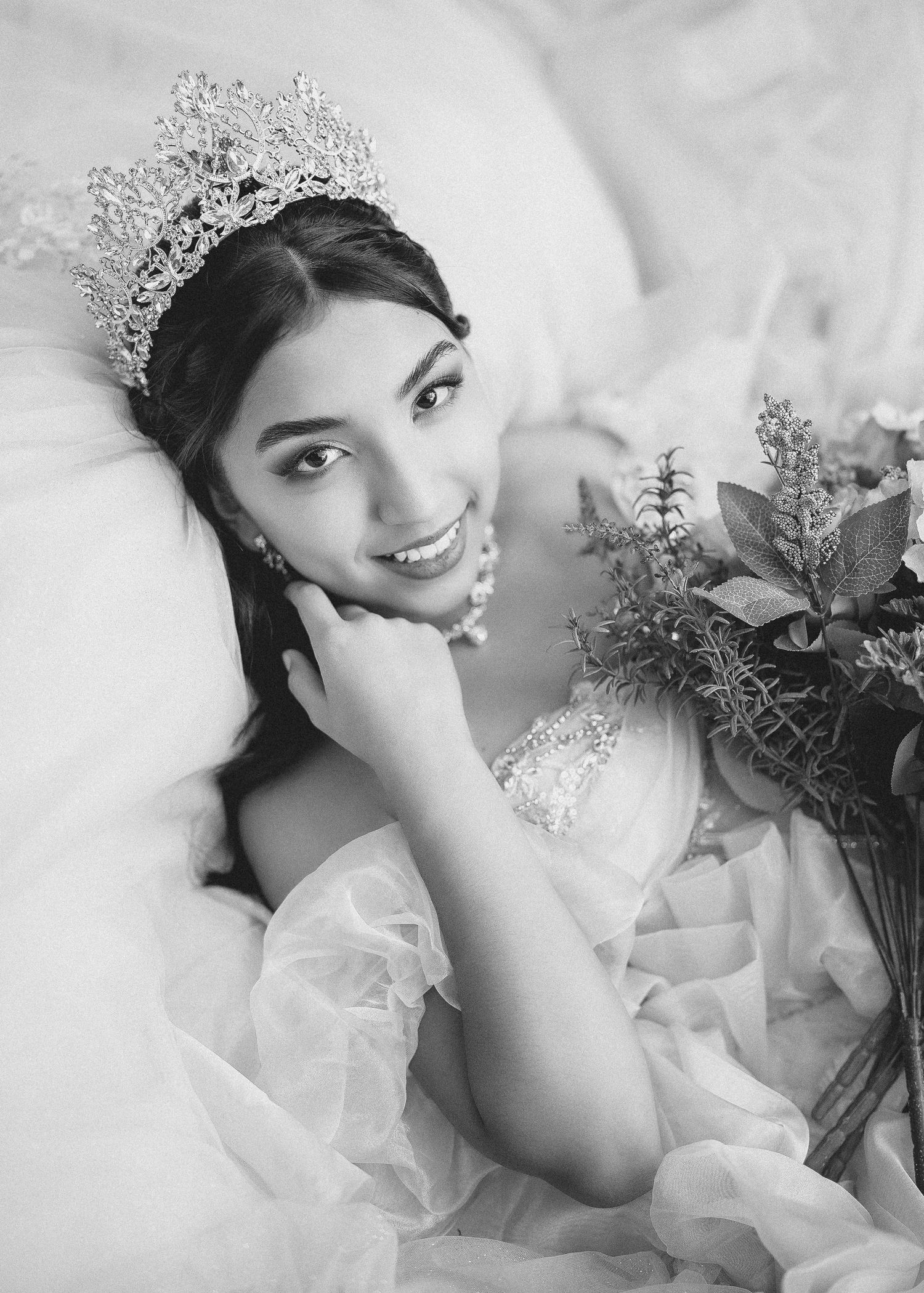 A bride in a white wedding dress lies on white bedding while holding a bouquet of flowers.