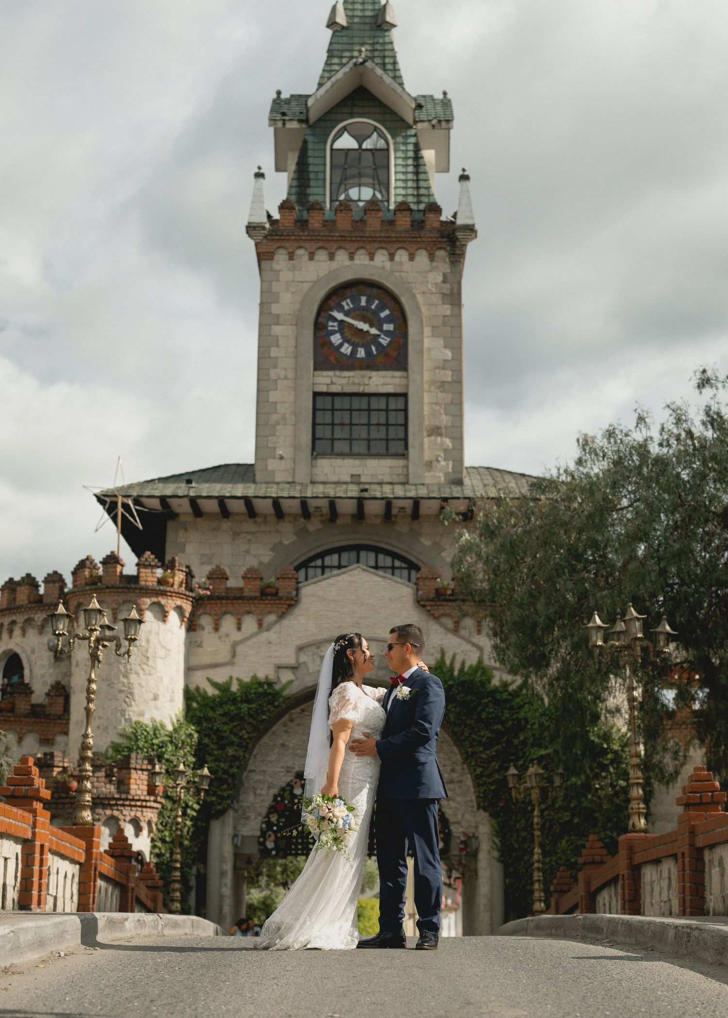 A couple stands in front of a historic clock tower with gothic architecture on their wedding day.