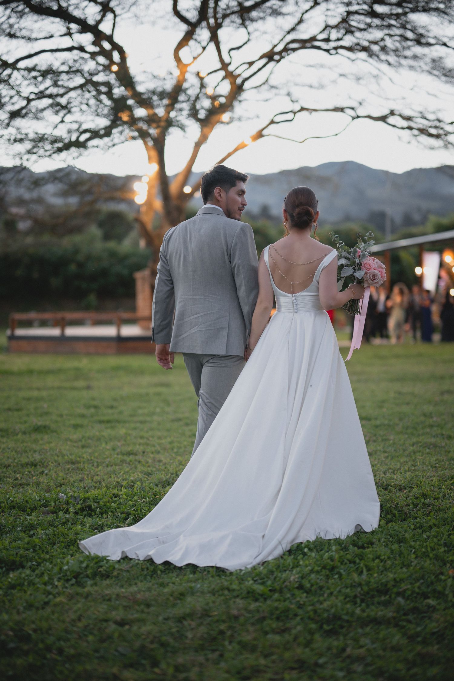 A couple in wedding attire walks together on a grassy field during sunset with trees in the background.