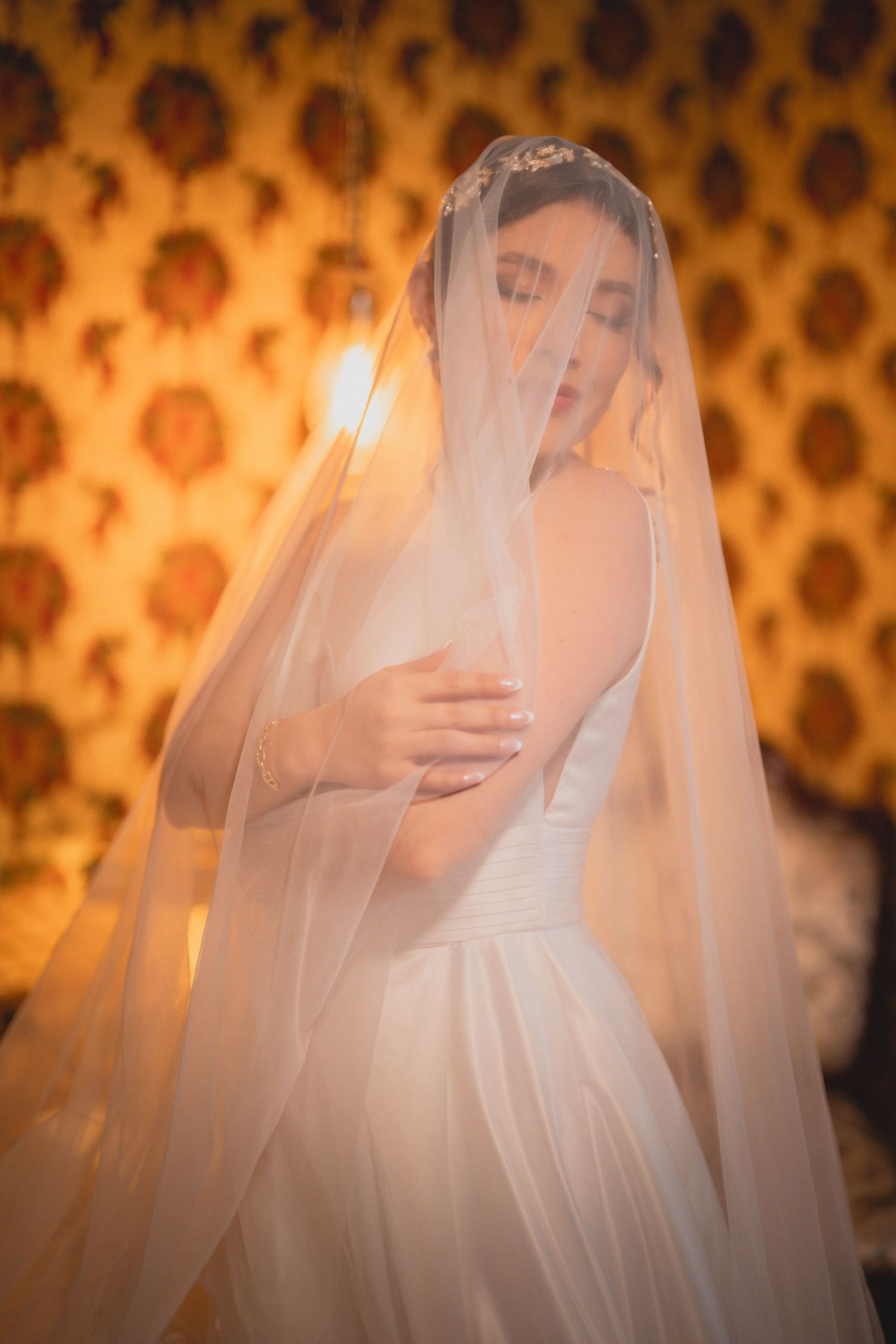 Ethereal bridal portrait with sheer veil against golden patterned wallpaper in warm ambient lighting.