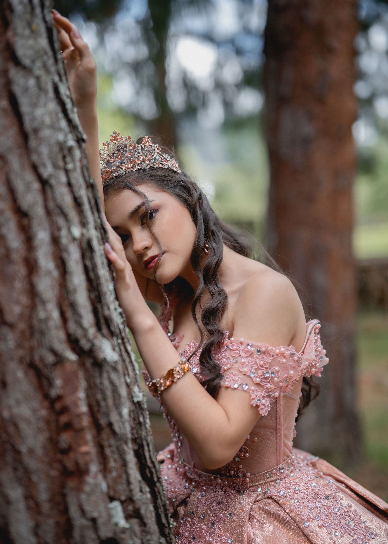 A person in a pink sequined gown poses elegantly against a tree trunk in a forest setting.