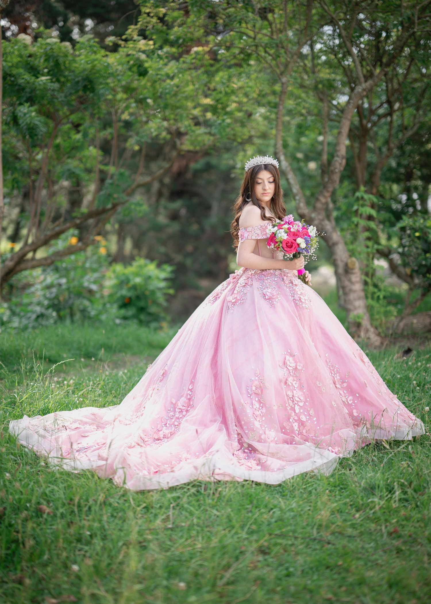 A flowing pink lace ballgown dress spreads elegantly across green grass in an outdoor garden setting.
