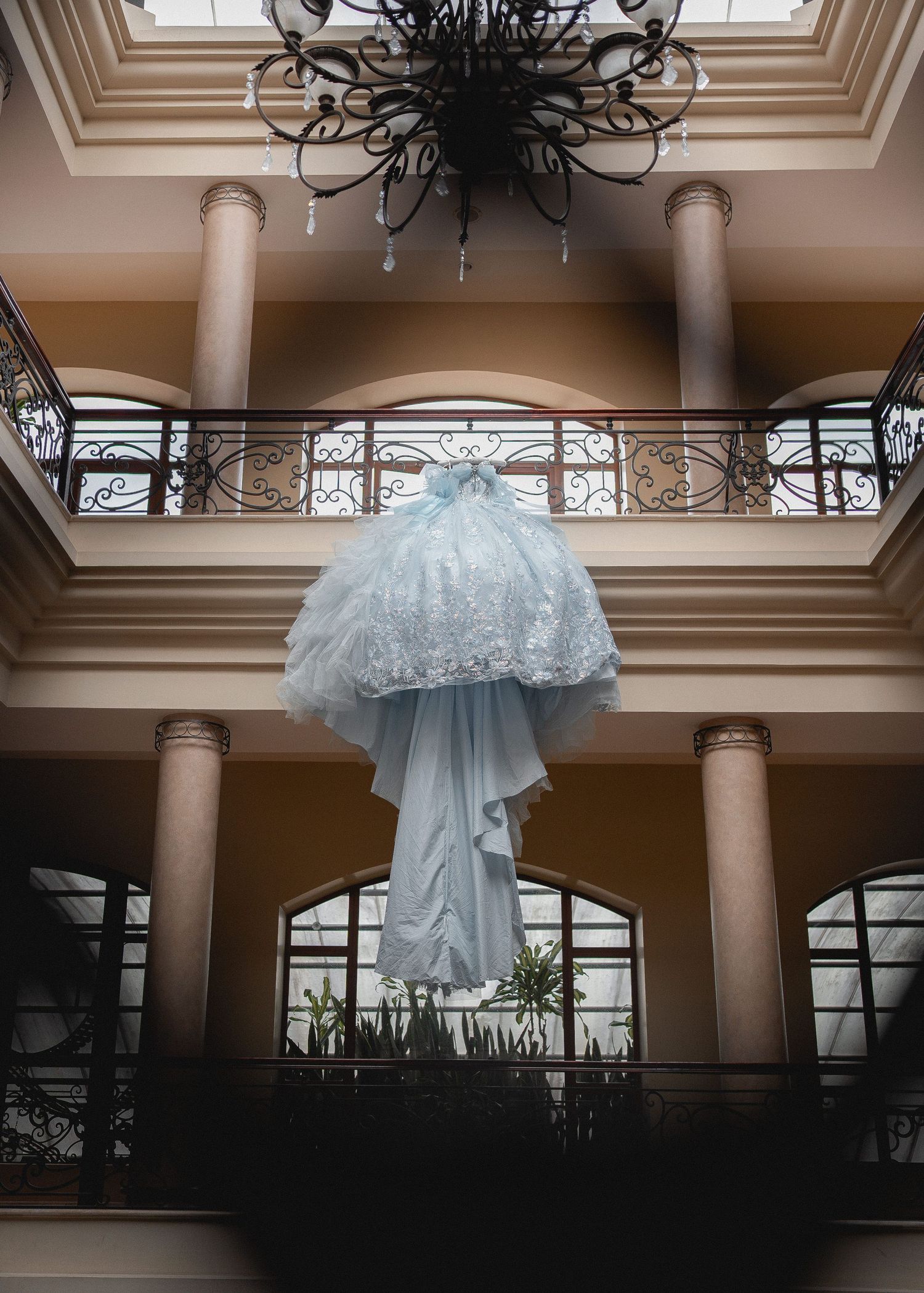 A series of light blue tulle wedding dresses hang from an ornate ceiling in an elegant ballroom interior.