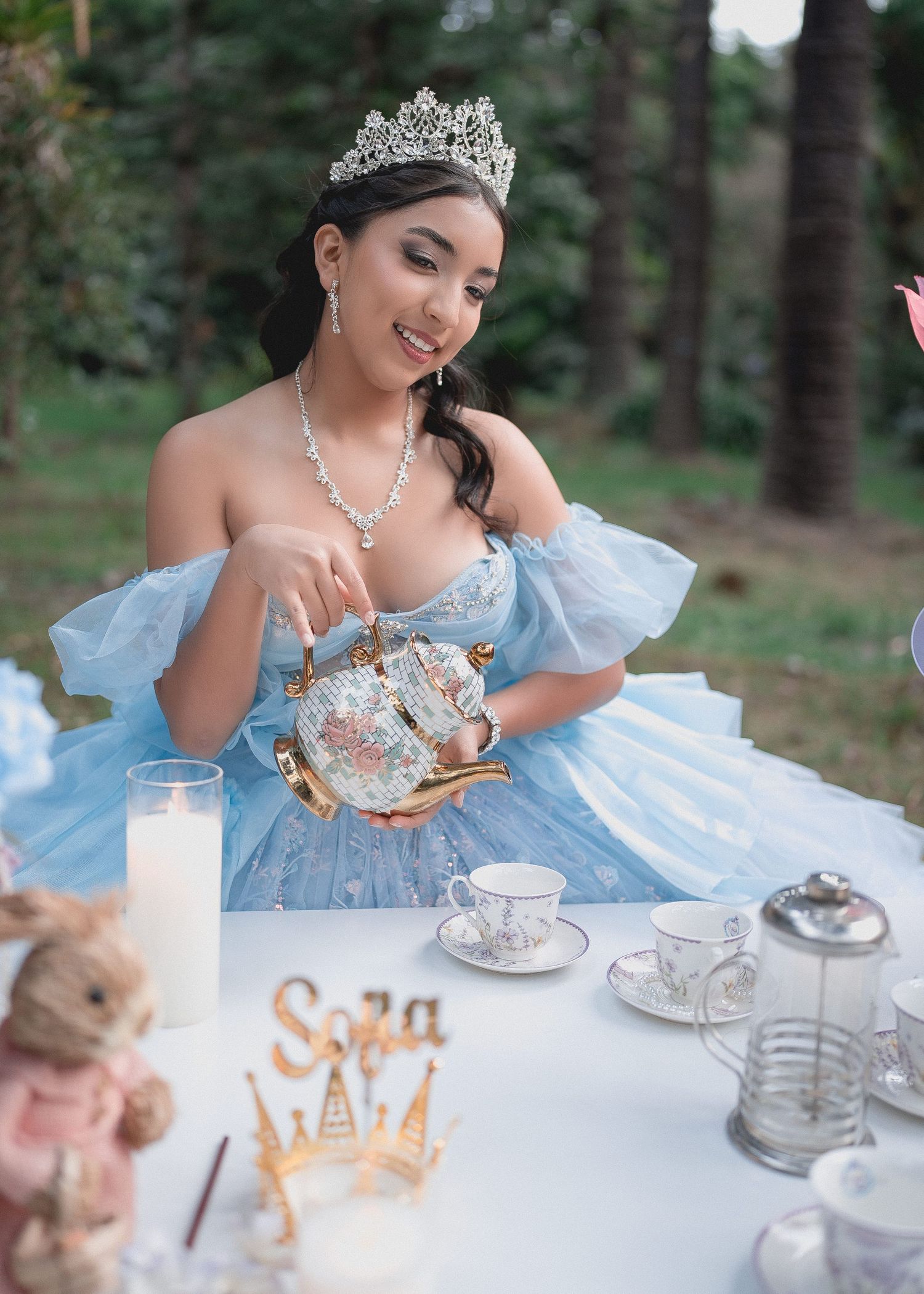A dreamy tea party setup in the woods with a light blue gown, vintage teacups, and delicate pastries on a white tablecloth.