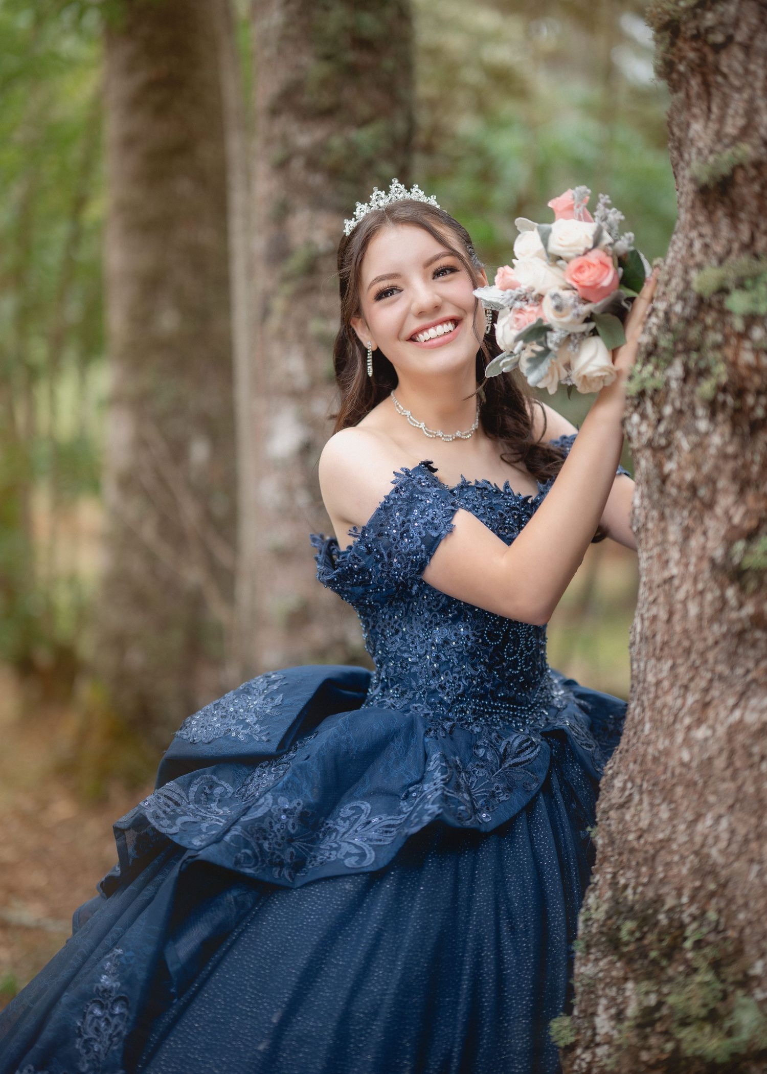 A person in an elegant navy blue lace ball gown poses in a forest setting while holding a pink and white bouquet.