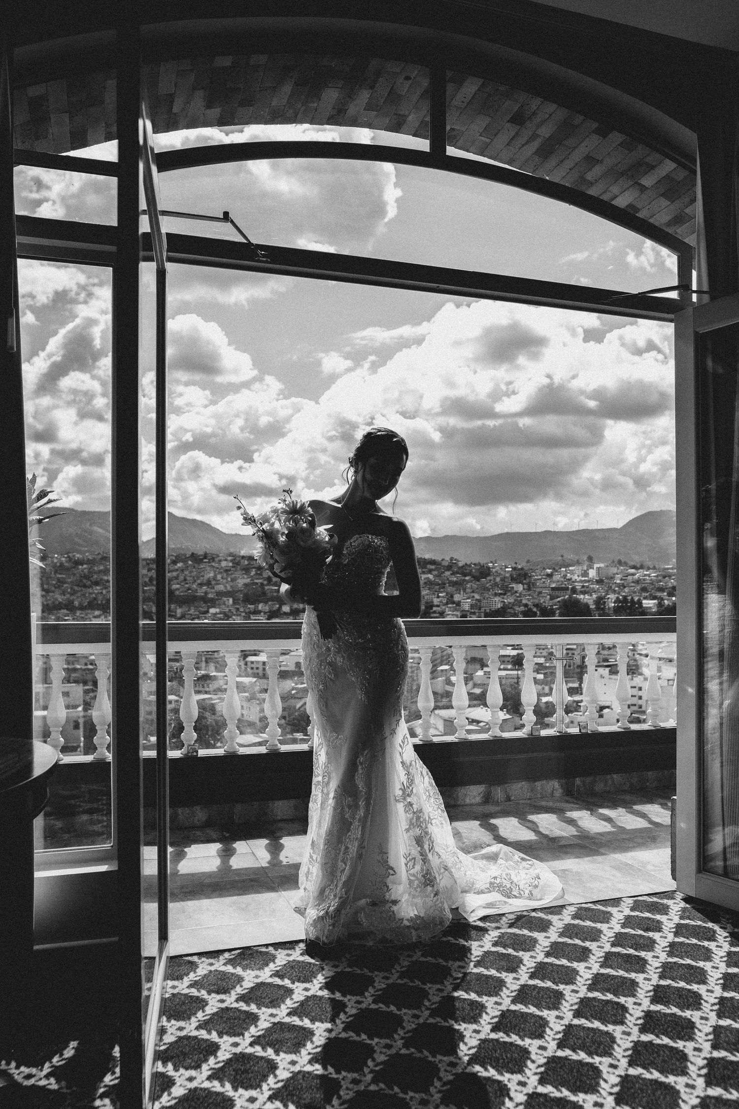 A silhouette stands on a balcony overlooking a scenic mountain landscape, holding flowers in a lace dress.