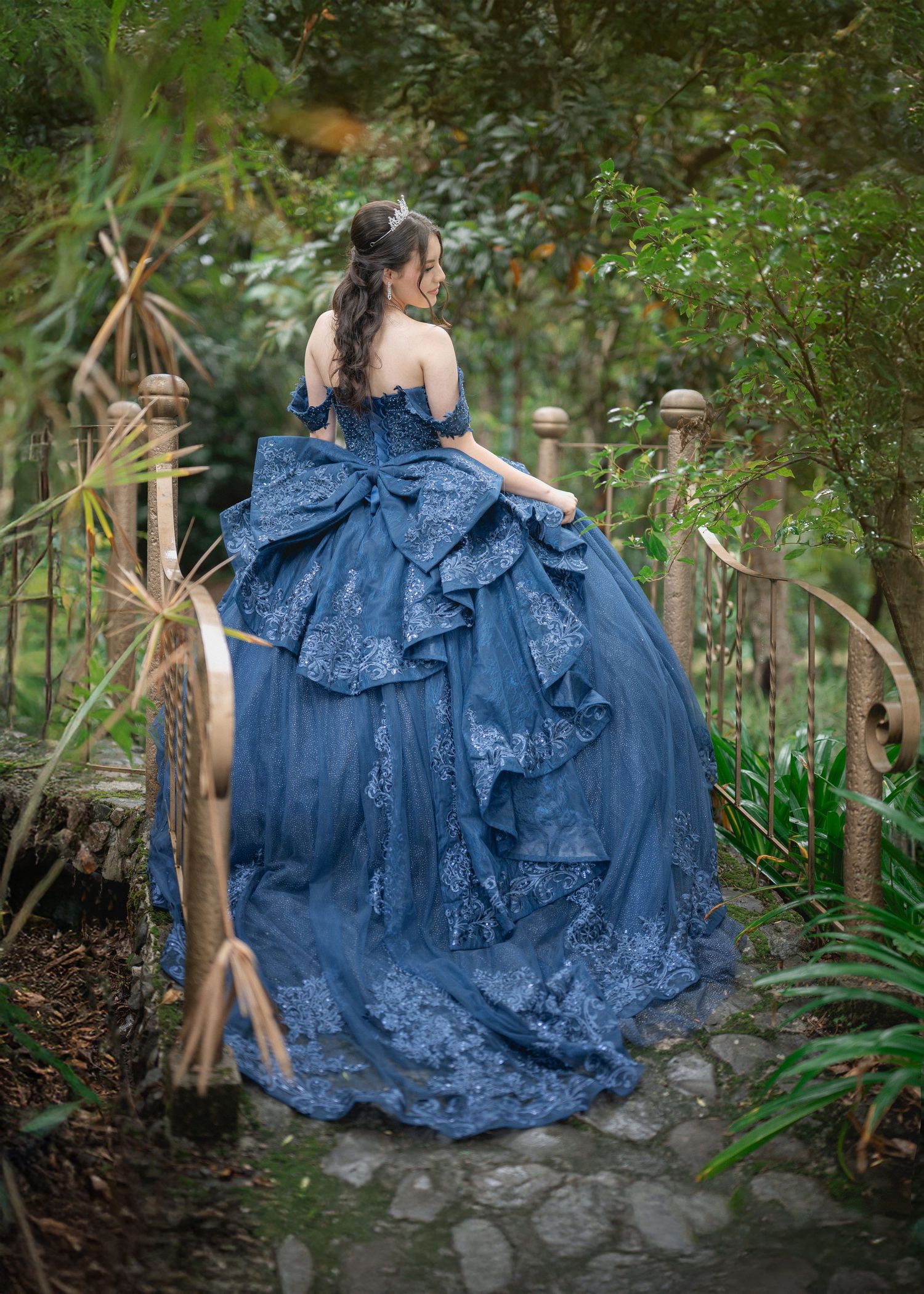 A person in a flowing blue vintage-style dress poses gracefully in a lush garden setting with stone path details.