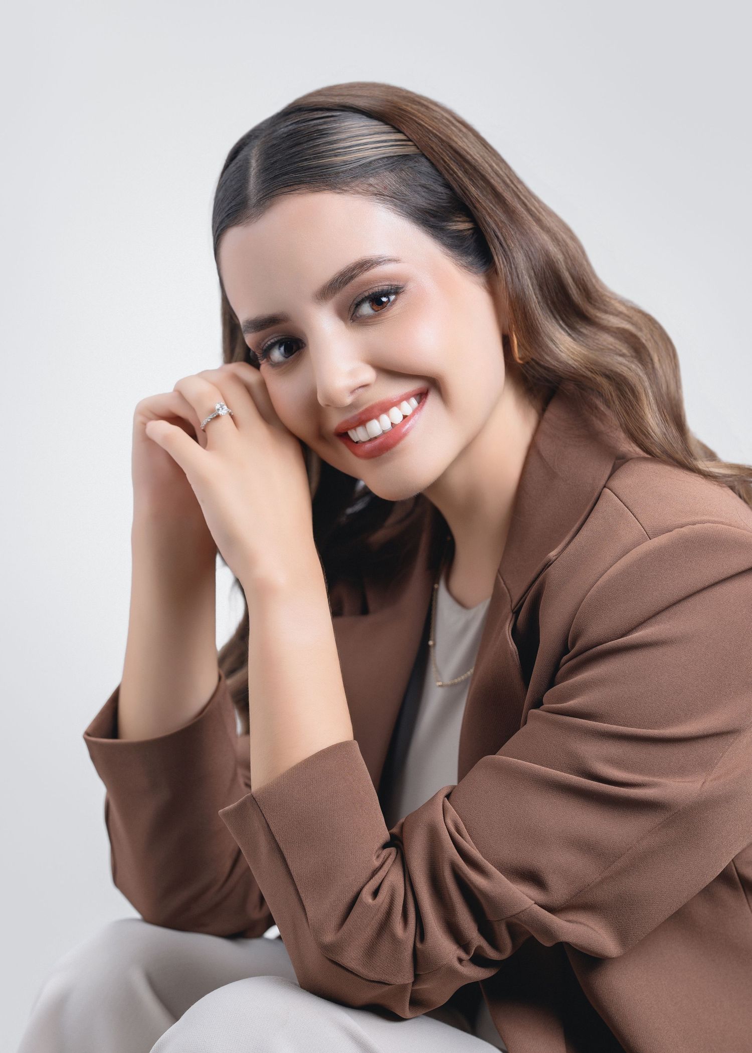 Professional portrait of someone wearing a brown blazer and white top while smiling warmly against a light background.