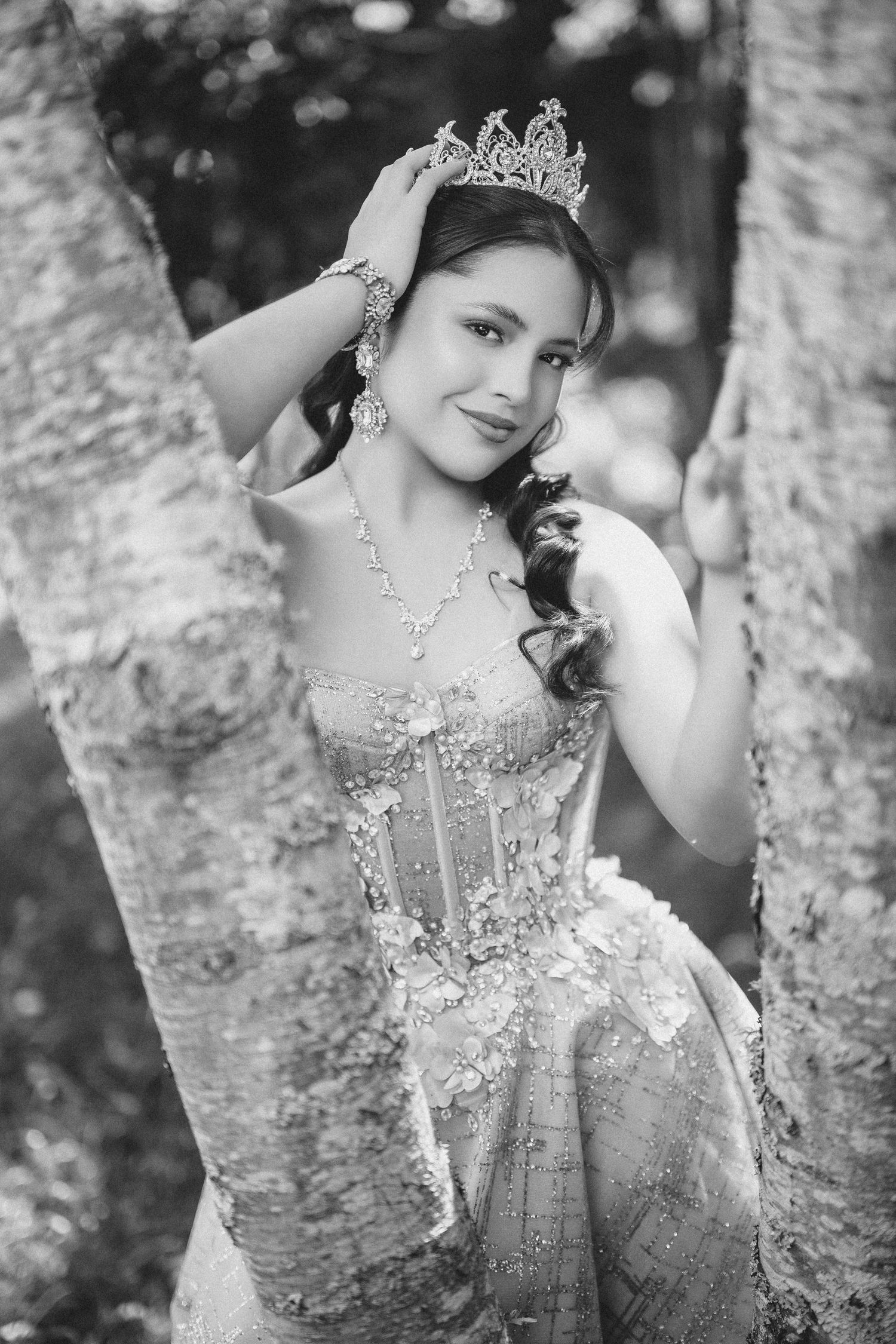 A monochrome photo of someone wearing an ornate ball gown and tiara while posing elegantly between trees.
