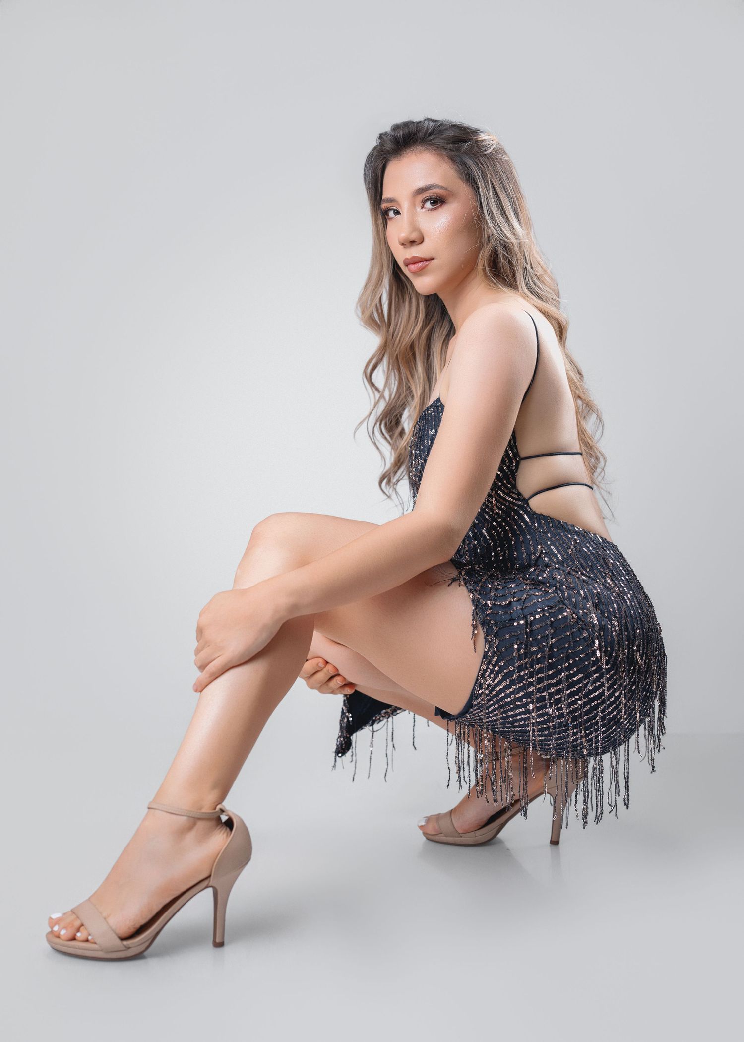 A model in a black sequined dress with fringe detail poses elegantly on a light gray studio backdrop.