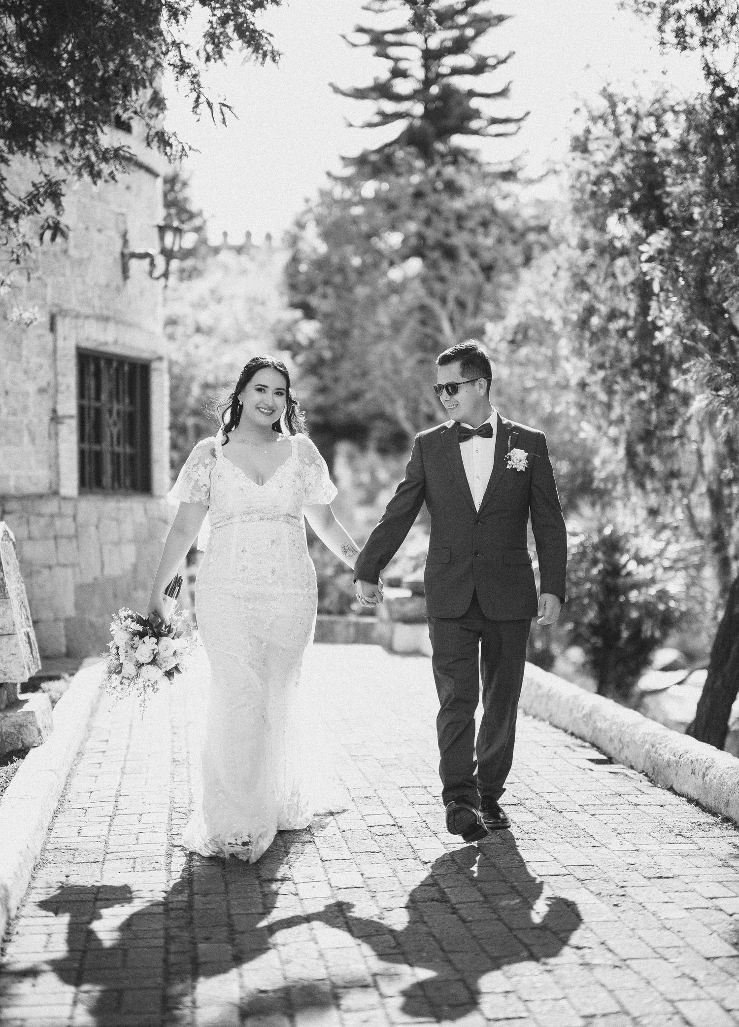 A romantic black and white wedding photo series showing a couple walking along a stone pathway surrounded by trees.