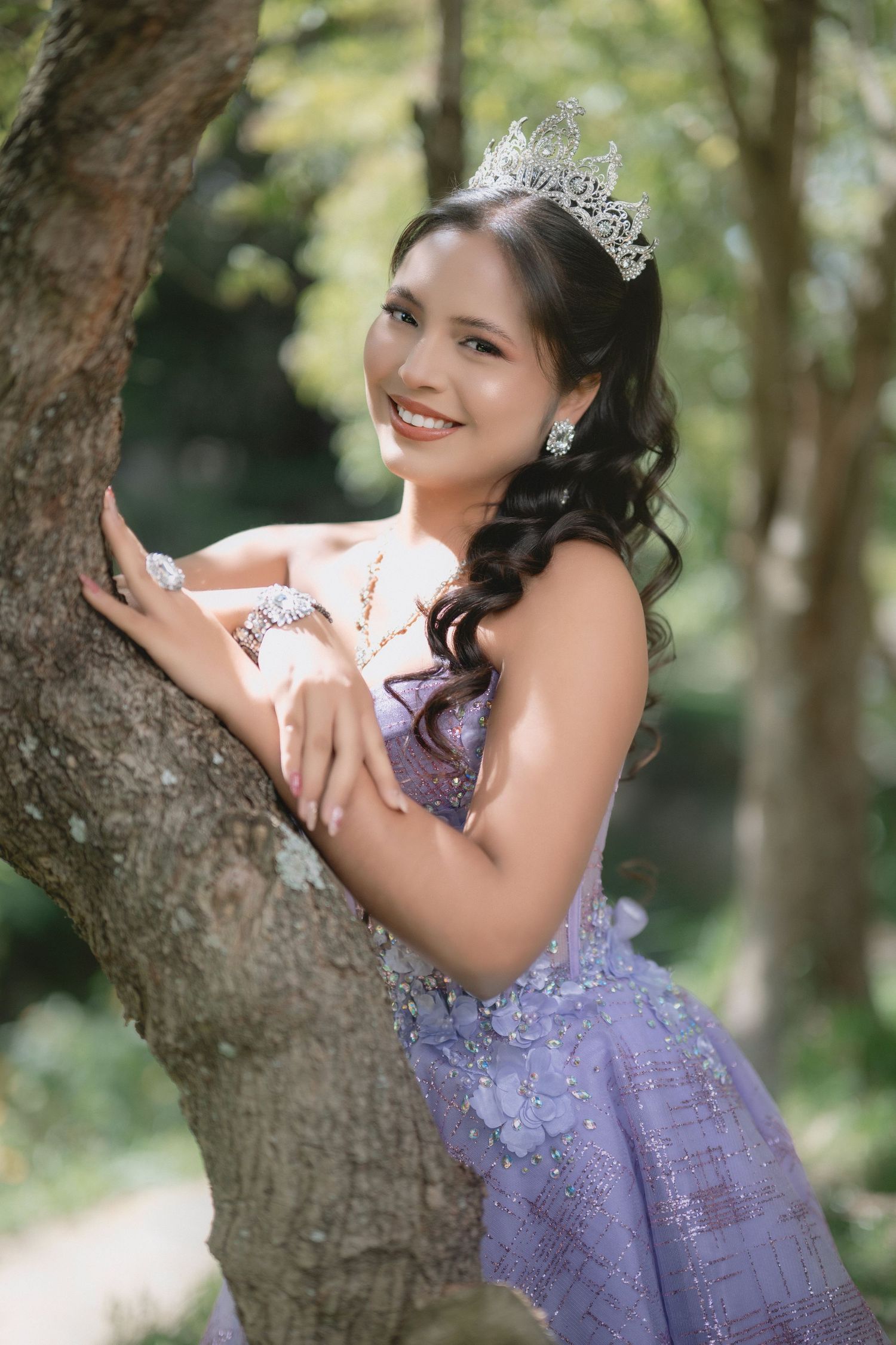 A person in a lavender gown and sparkly tiara poses elegantly against a tree trunk in a dreamy outdoor setting.