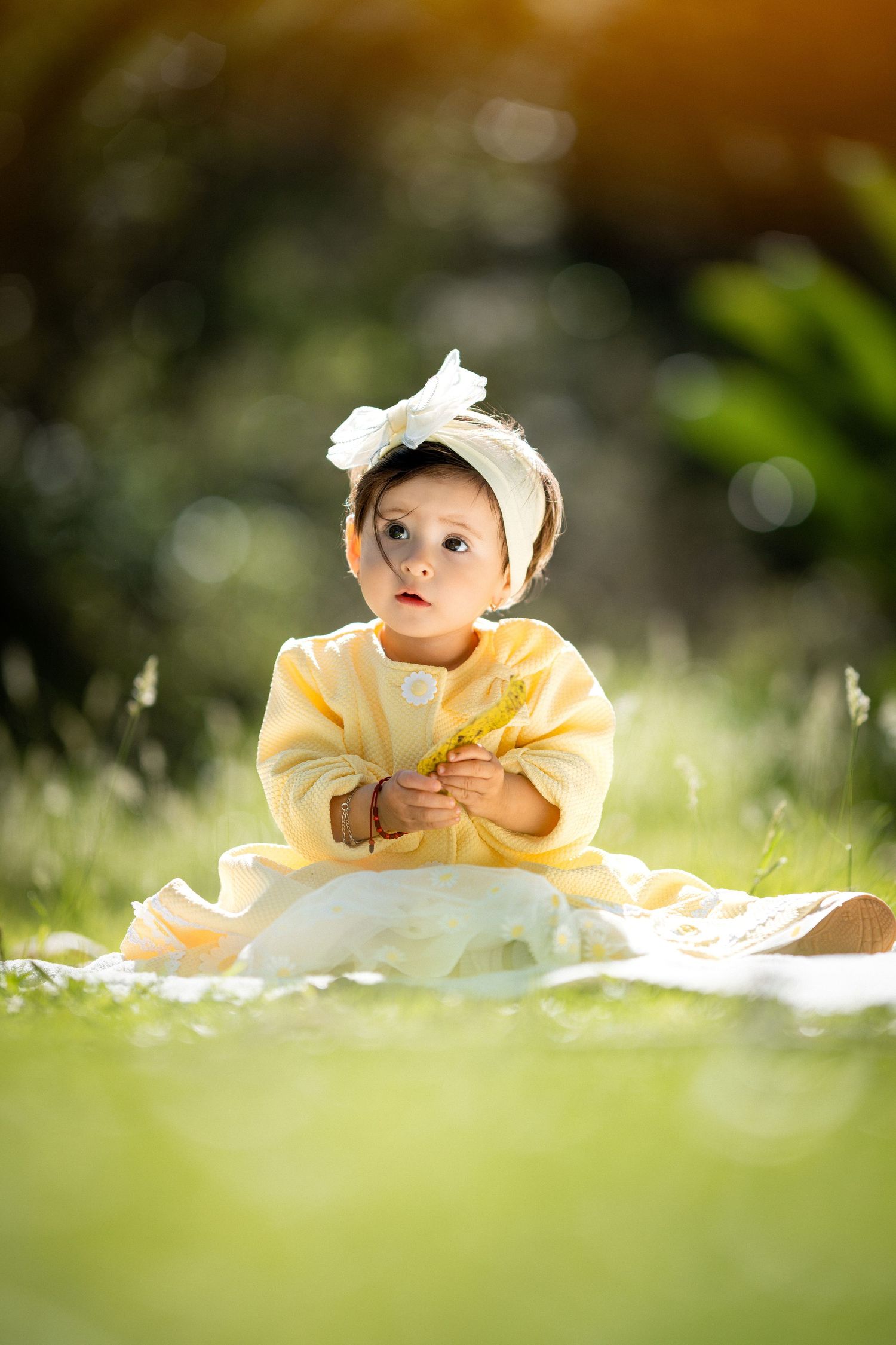 A small baby in a yellow outfit lies on a white blanket in the grass on a sunny day.