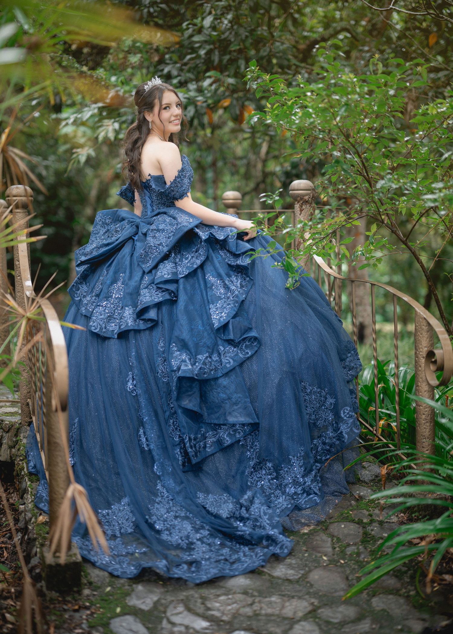 A woman in a flowing blue lace gown poses gracefully in a lush garden setting, creating a romantic outdoor portrait series.
