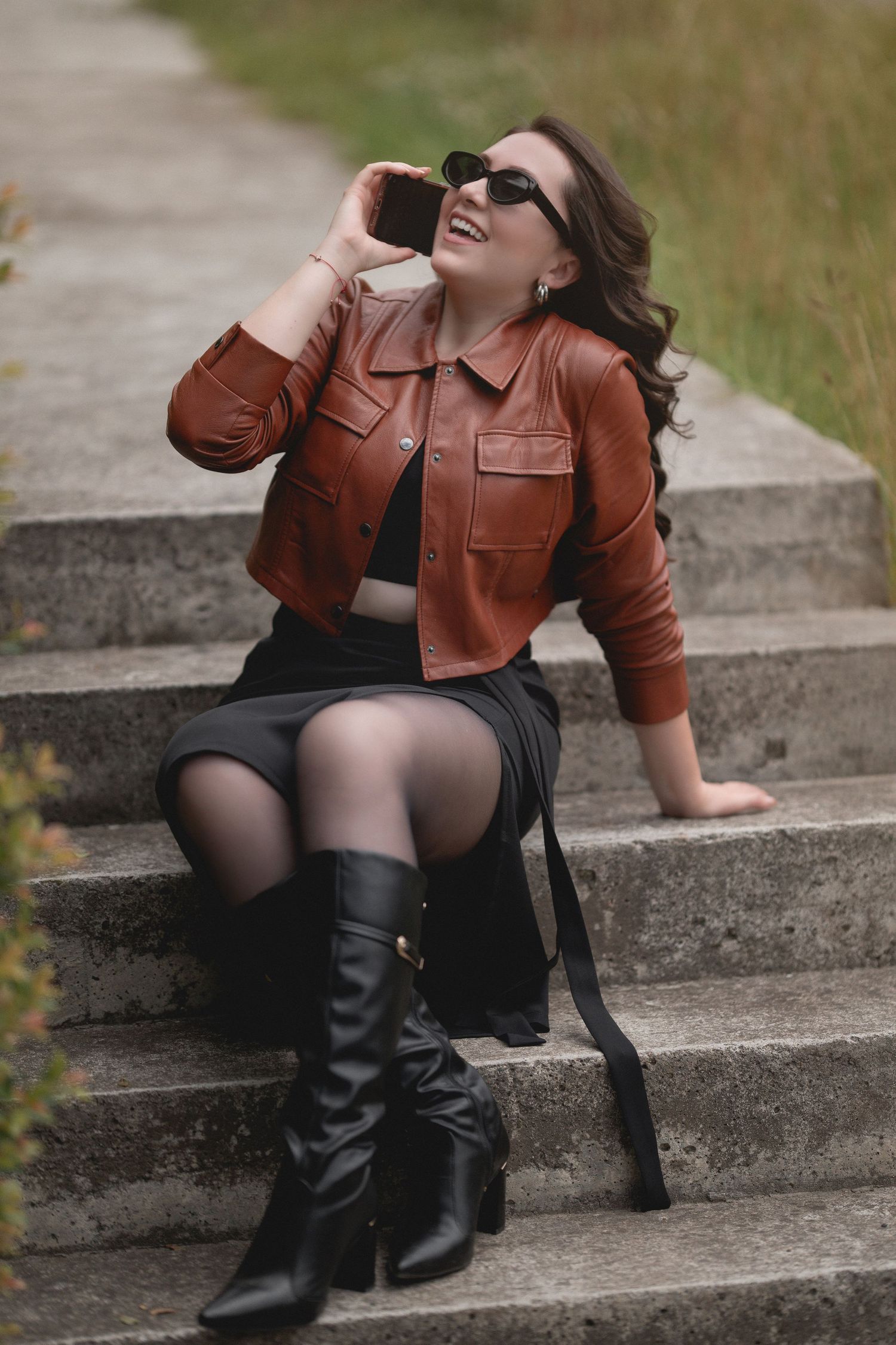 A person in a brown leather jacket and black boots poses dramatically on concrete steps while wearing sunglasses.