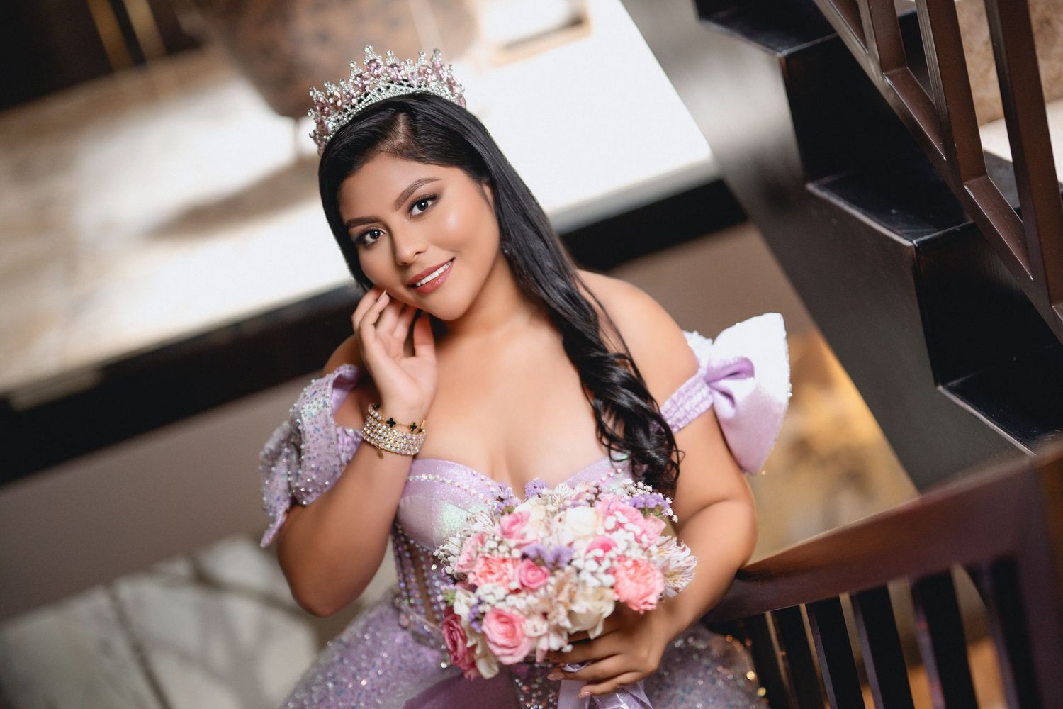 A woman in a lavender quinceanera dress poses elegantly on a wooden staircase with pink and white floral accents.