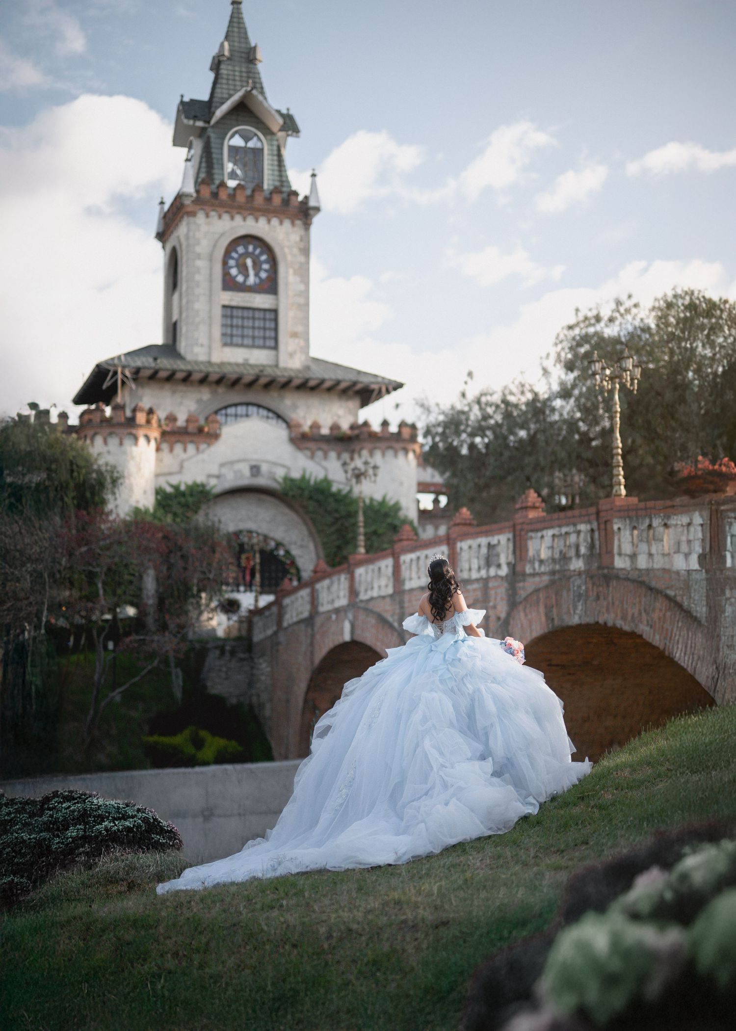 A wedding dress train flows gracefully along a grassy hill beside a stone castle tower with arched bridges at sunset.