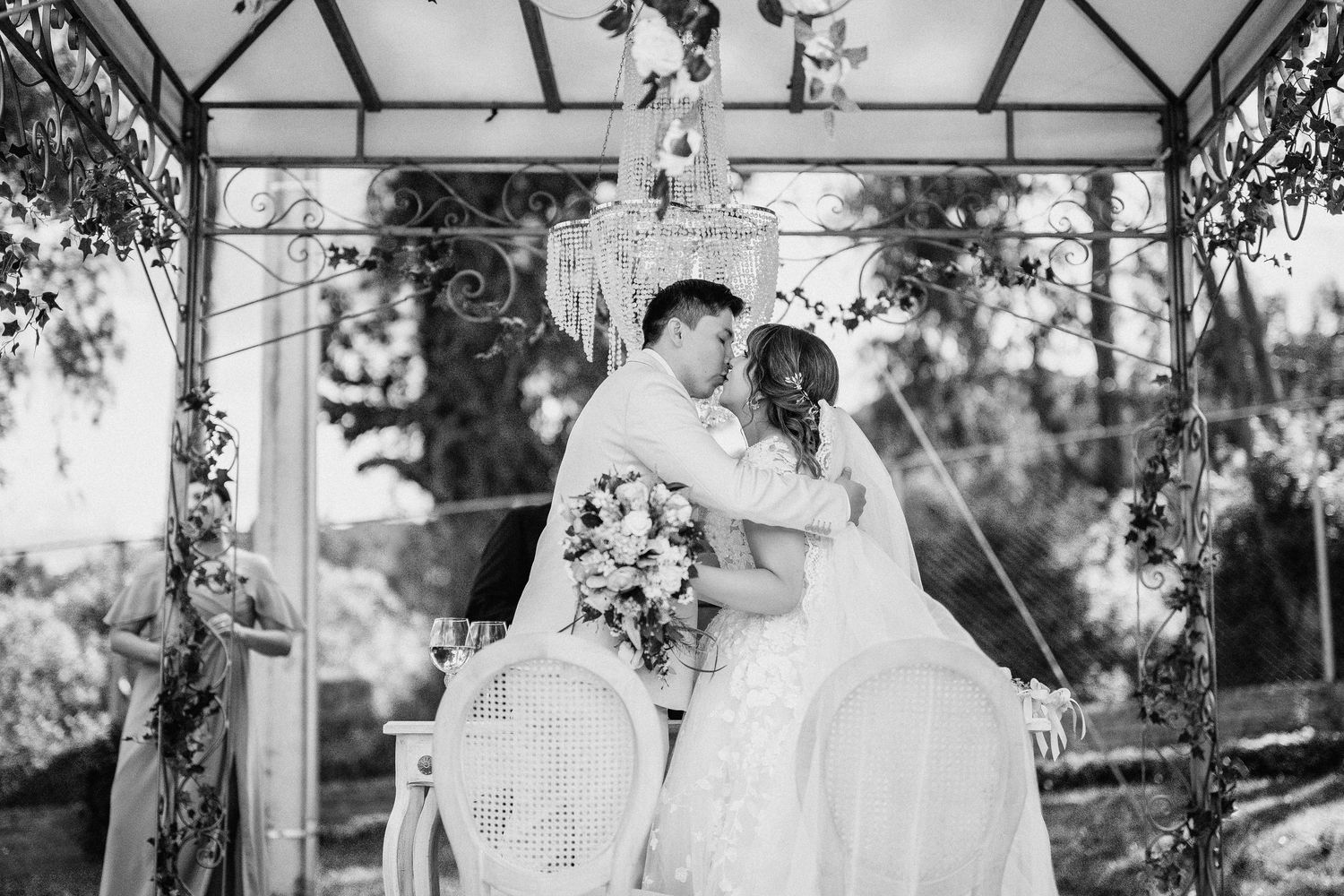 A romantic outdoor wedding ceremony with floral arrangements and guests seated in white chairs under a canopy of trees.