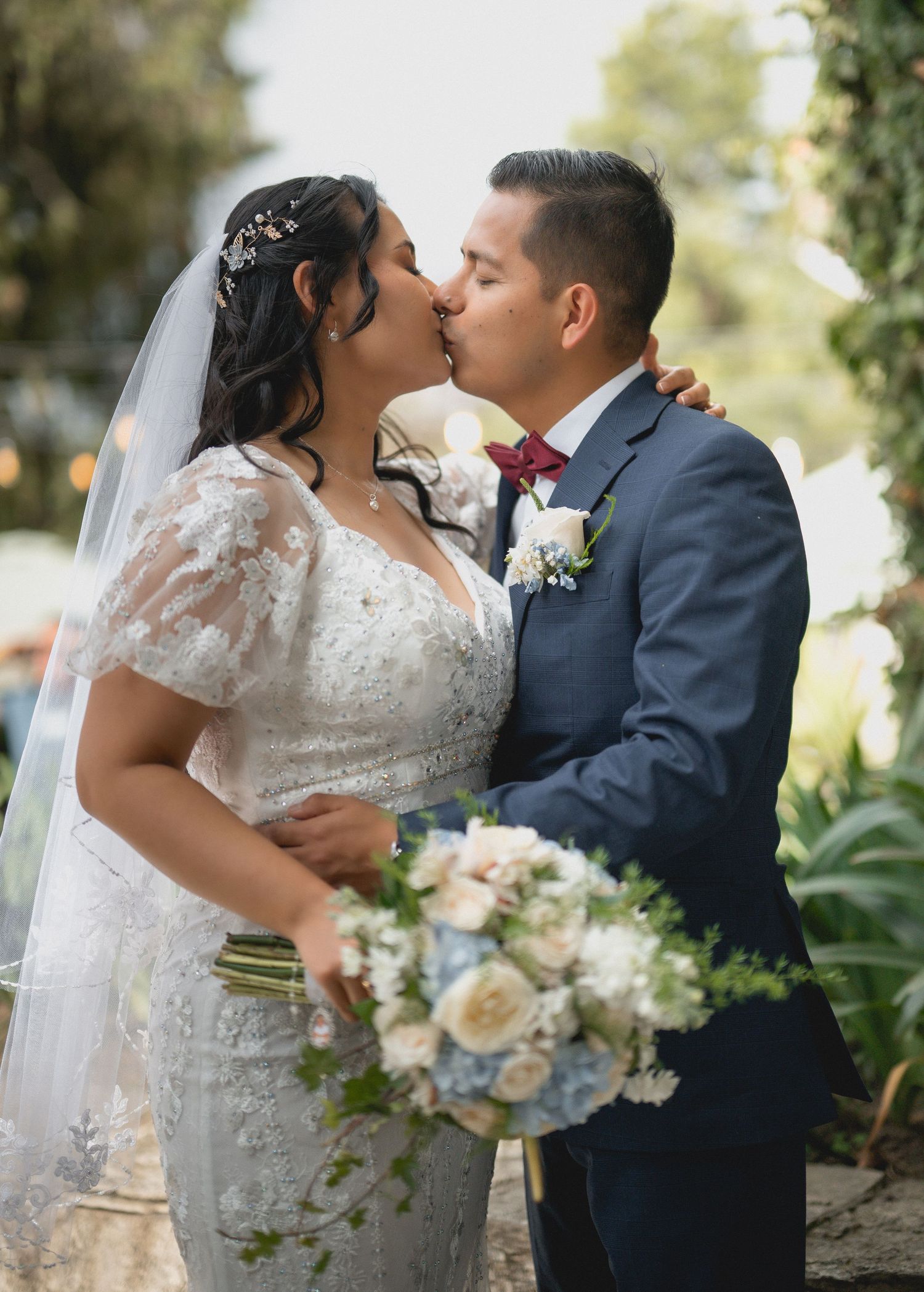 A romantic wedding couple shares intimate kisses while holding a bouquet of blue and white flowers in an outdoor garden setting.