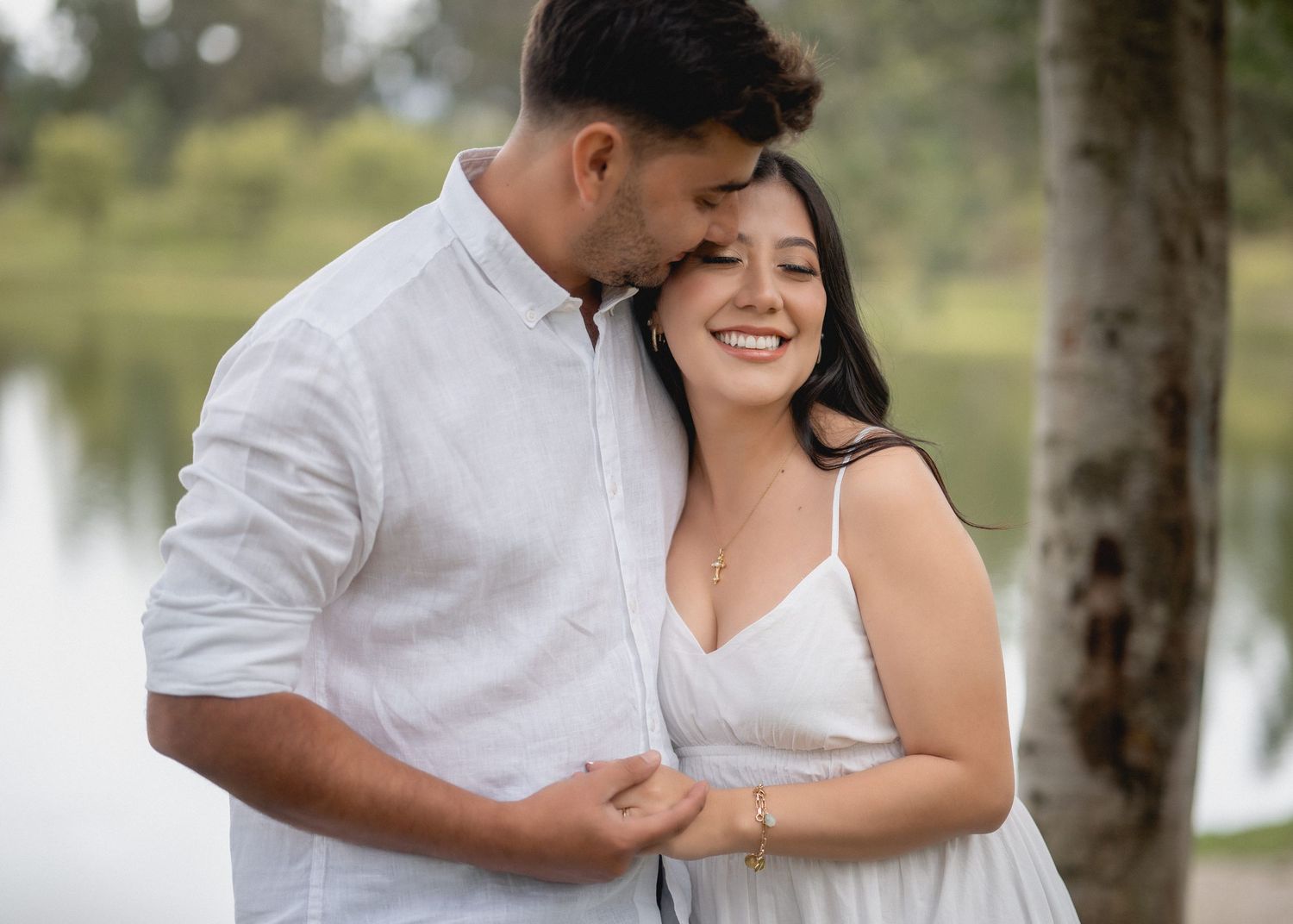 A romantic couple in white outfits share tender moments during an outdoor engagement photoshoot by a lake.