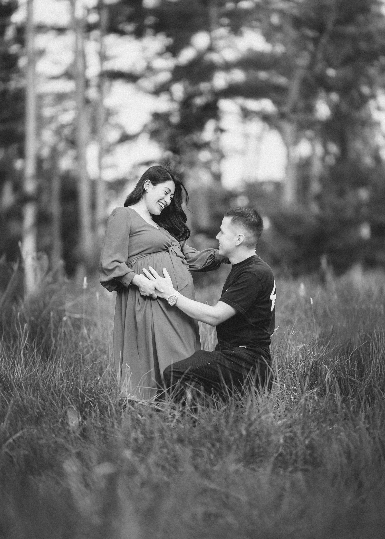 A romantic couple sits together in tall grass during an outdoor photoshoot in black and white.