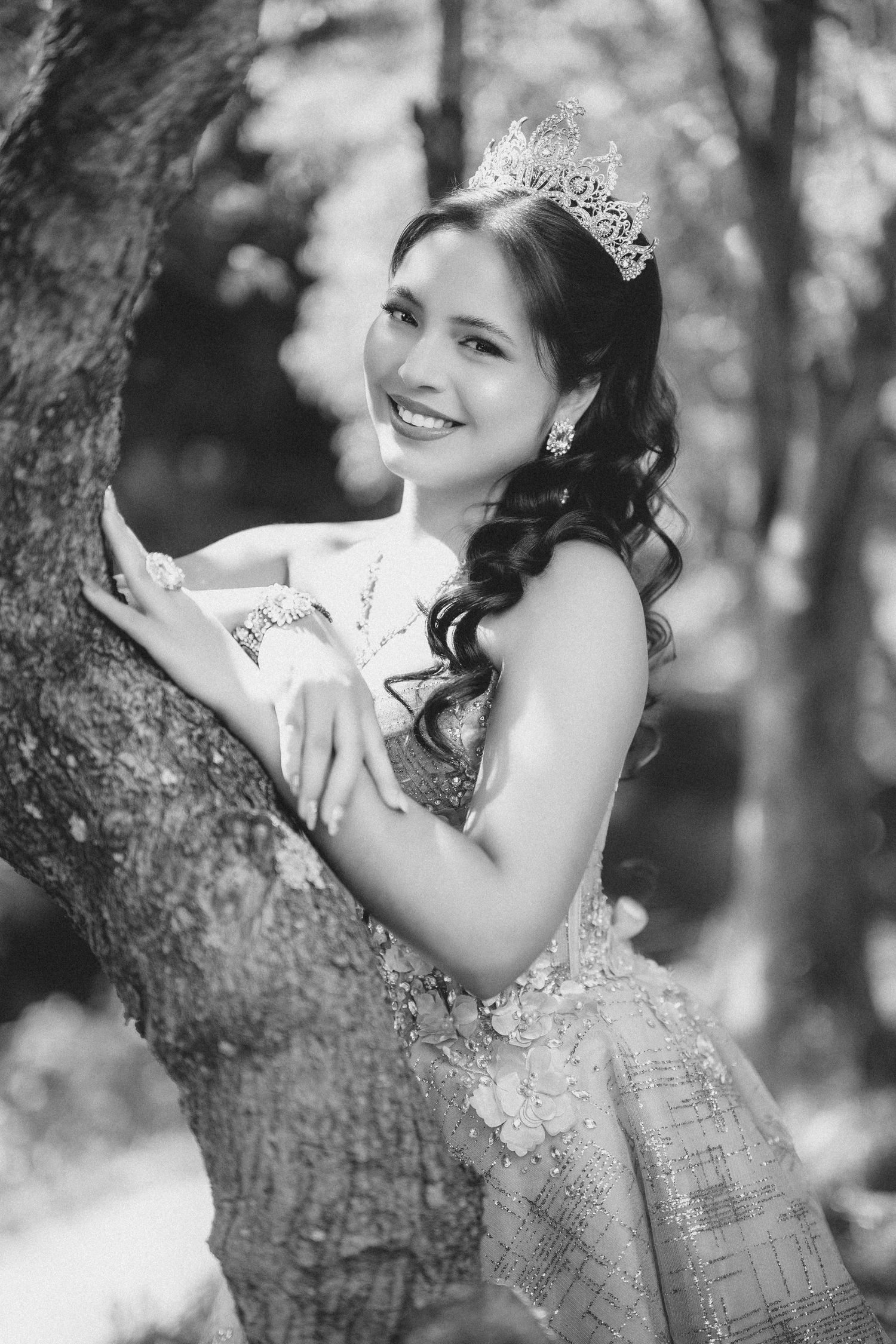 A black and white portrait of someone in a princess gown and tiara posing against a tree in a dreamy forest setting.