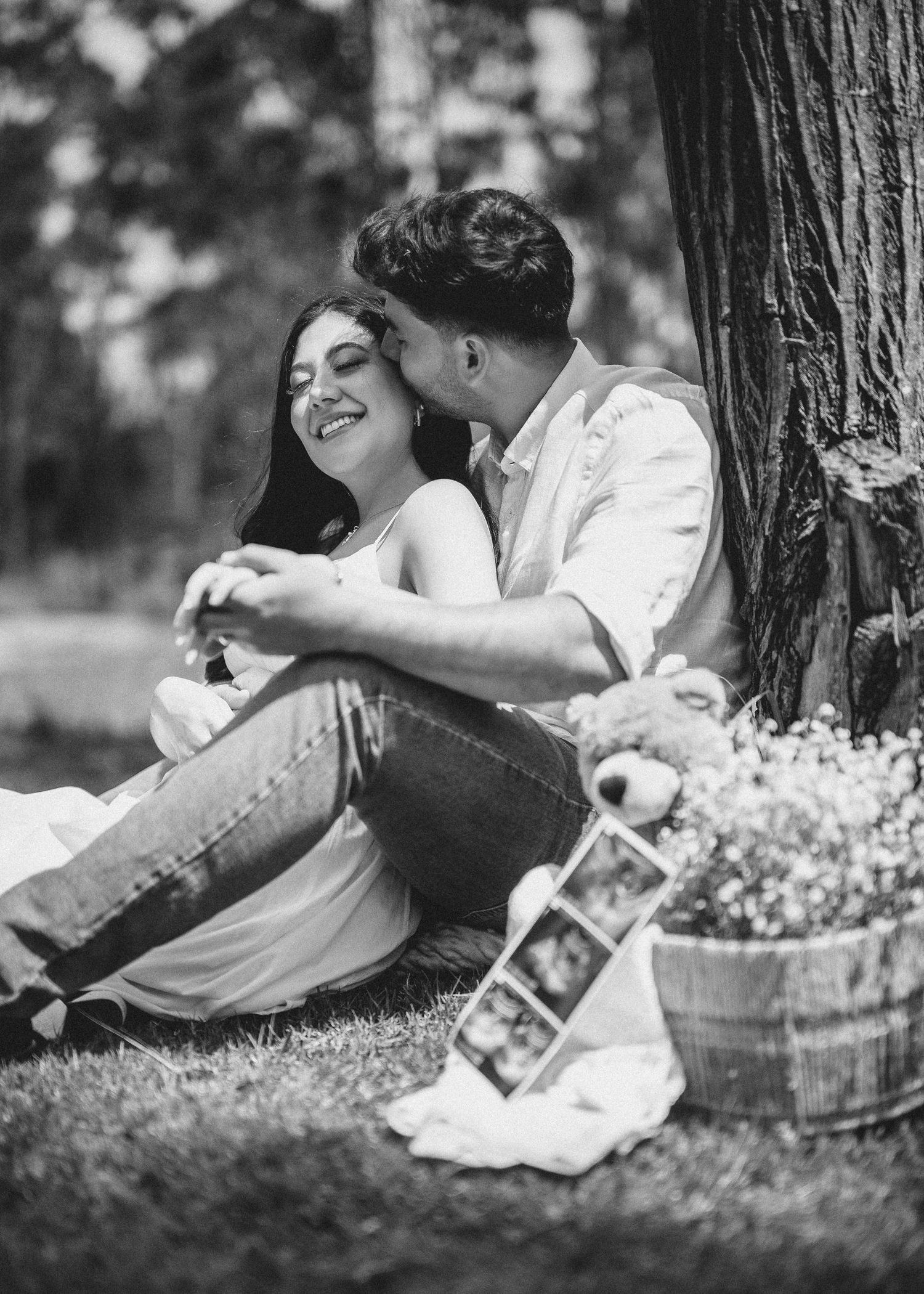 A romantic black and white photo of a couple sharing a tender moment while sitting against a tree in a garden setting.