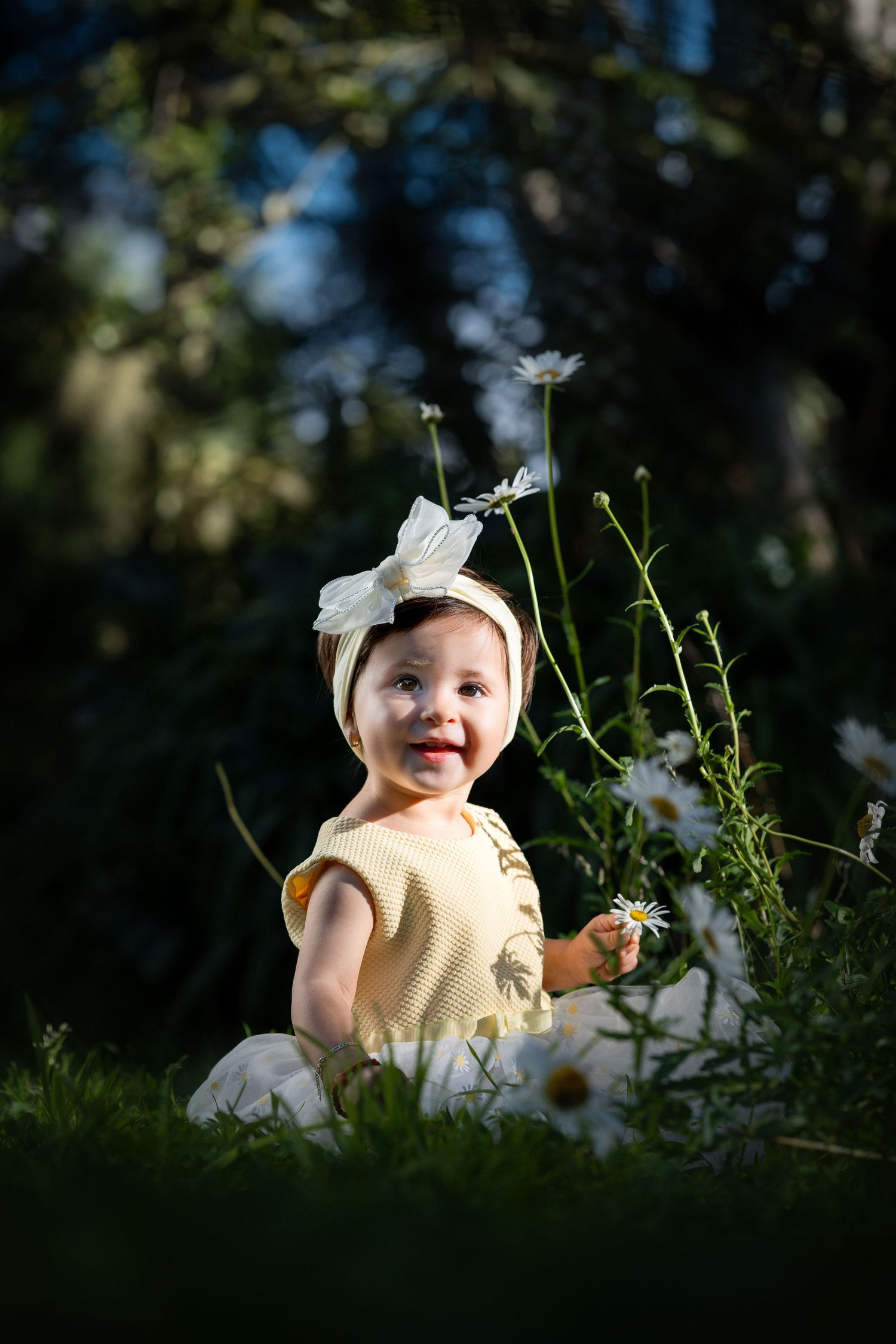 Baby in a white bow and yellow dress sitting among white wildflowers in dappled sunlight.