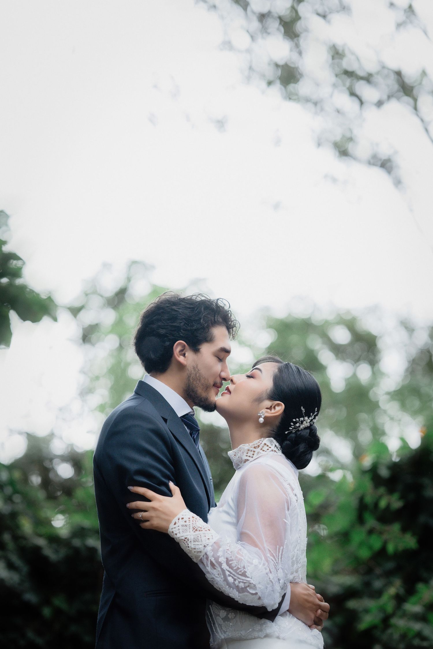 Romantic wedding couple embraces outdoors under trees in black and white attire.