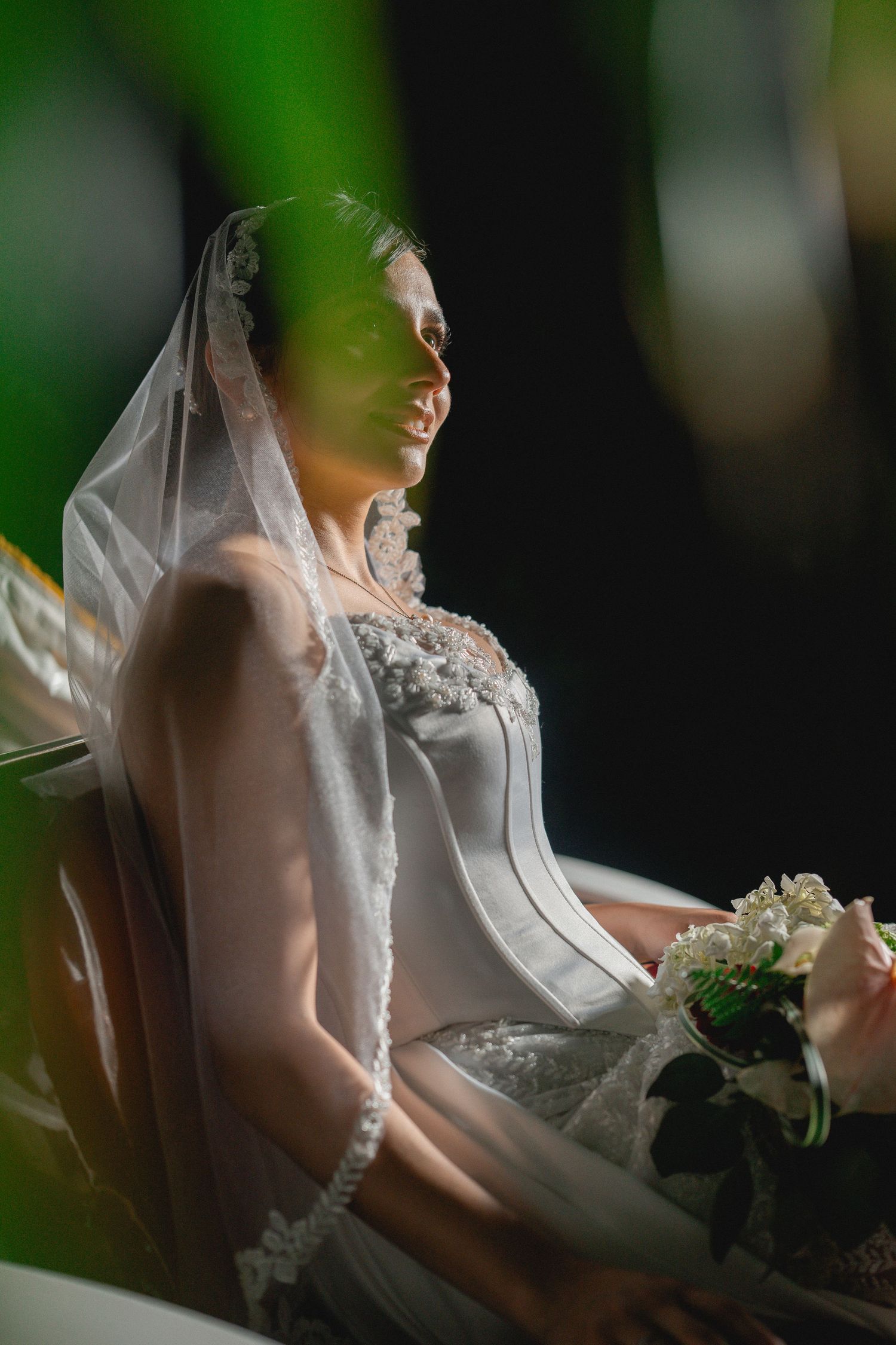 A bride in a white lace wedding dress and veil sits elegantly holding a bouquet with soft green bokeh in the background.