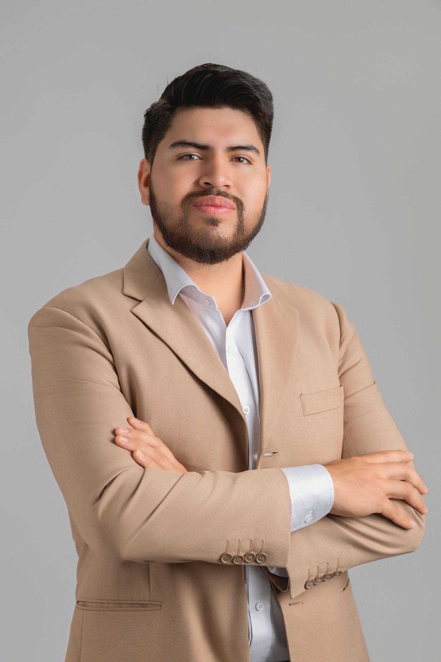 Professional wearing a tan blazer and white dress shirt poses with crossed arms against a gray background.