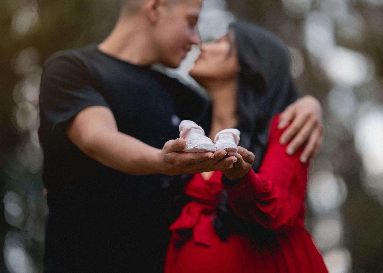 A romantic couple in a red dress and dark suit share an intimate dance pose outdoors in a forest setting.