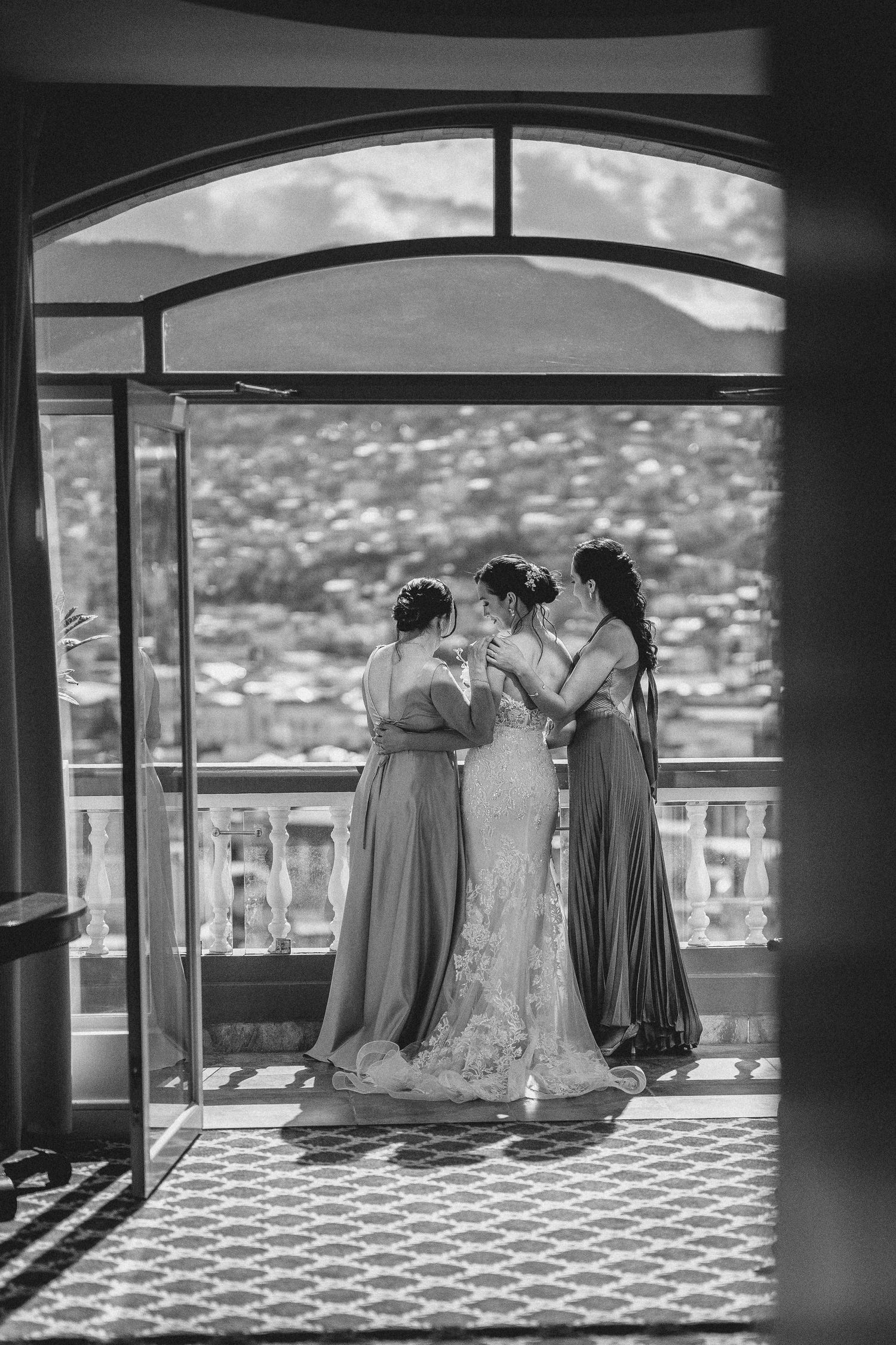 A bride and bridesmaids share an intimate moment on a balcony overlooking a scenic mountain landscape.