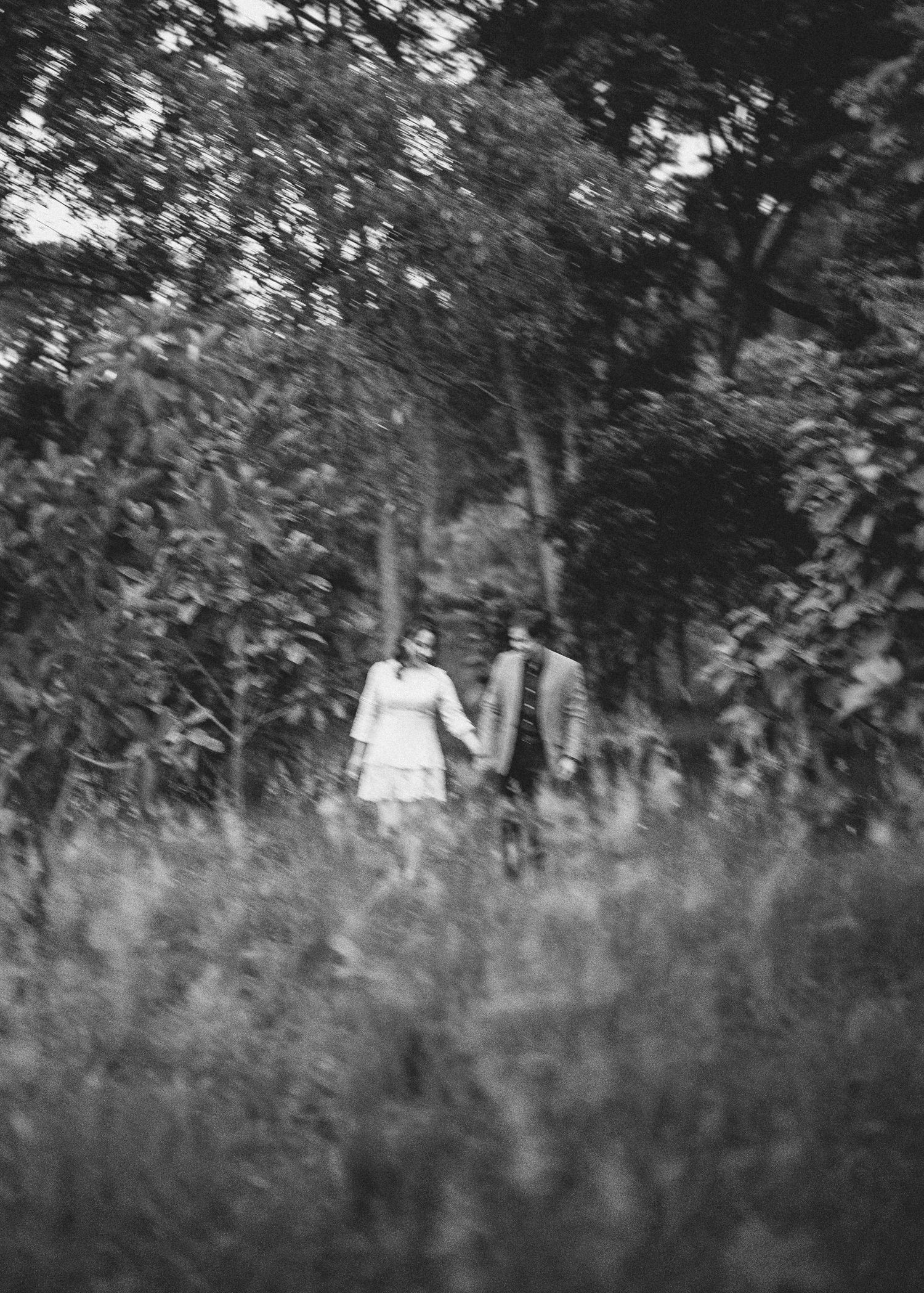 Black and white photograph showing a person walking through tall grass in a wooded area.