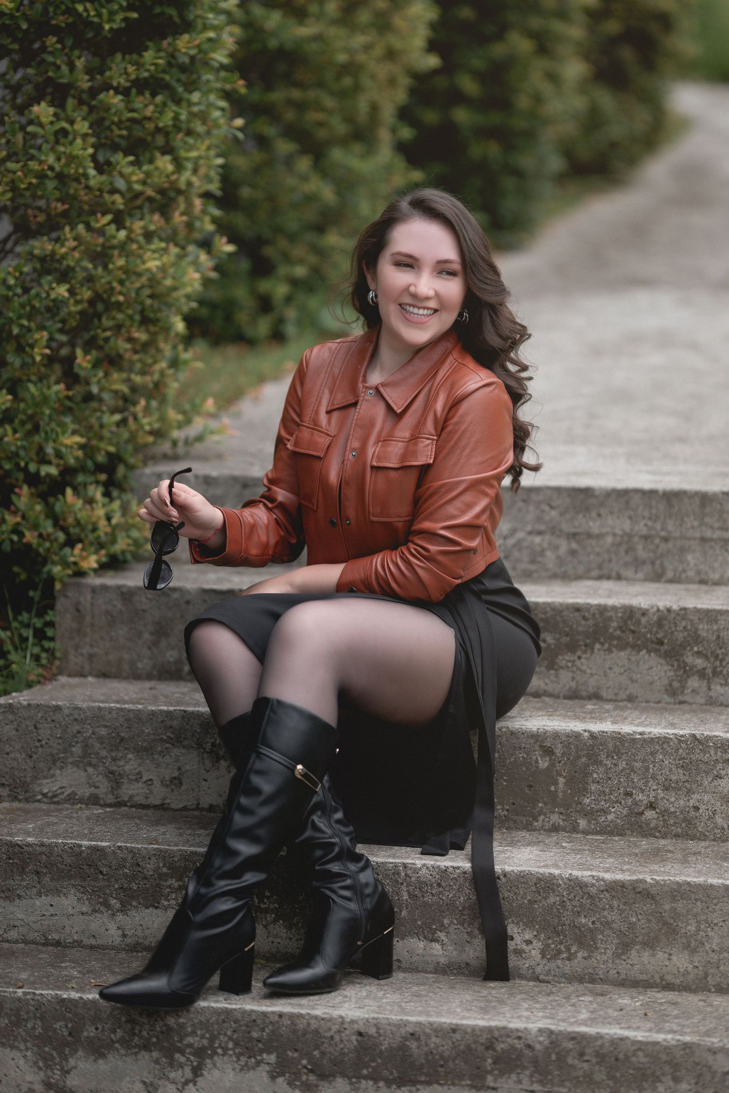 A person in a brown leather jacket and black boots sits on outdoor stone steps near green bushes.