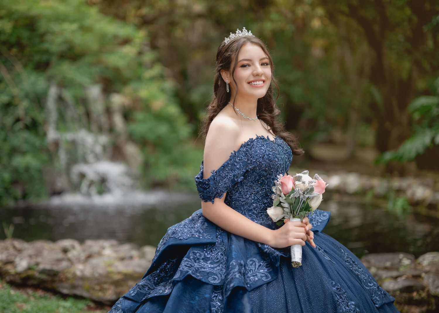 A woman in a navy blue off-shoulder ball gown holds a bouquet while posing by a waterfall in a garden setting.