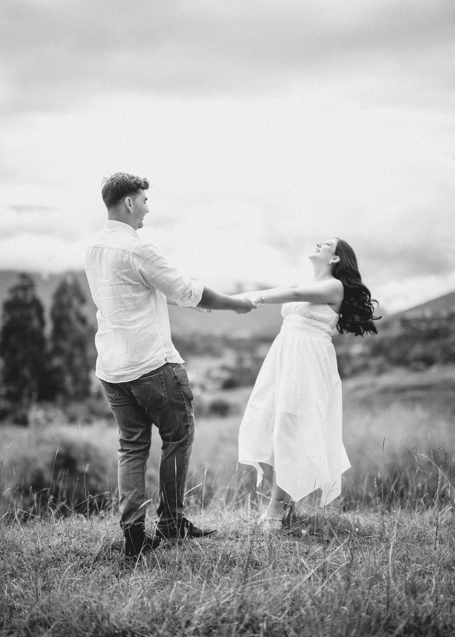 Black and white panoramic photo of a couple walking together through a grassy meadow on a cloudy day.