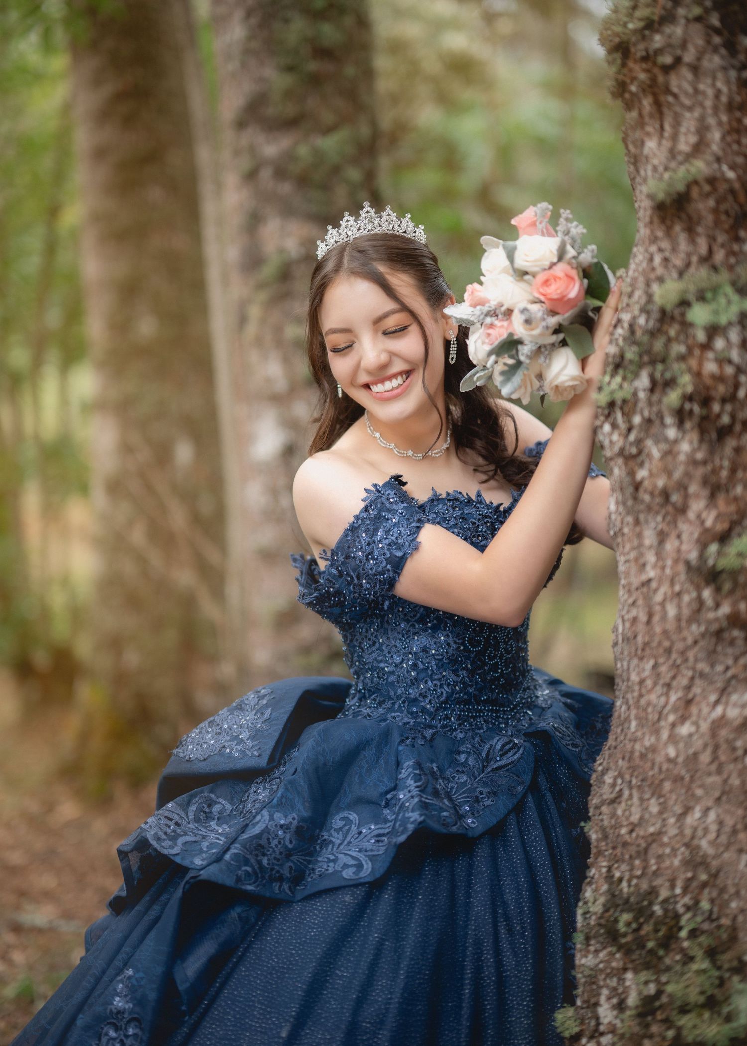 A person in a navy blue lace formal gown poses gracefully against trees in an outdoor forest setting.