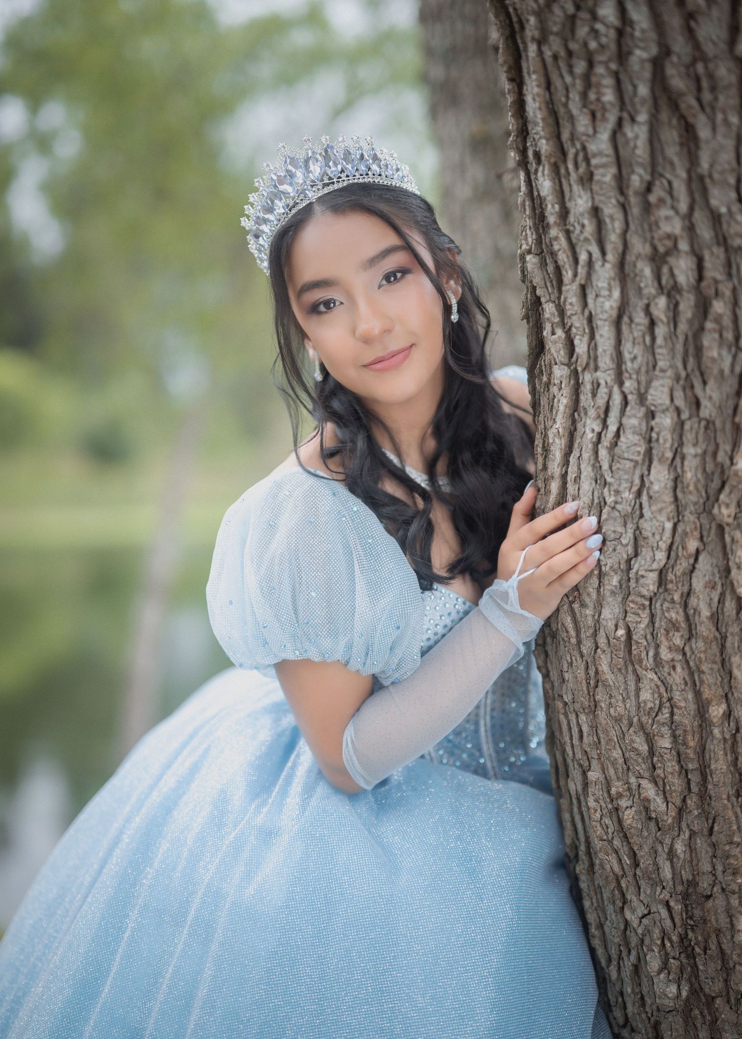 A person in a light blue princess dress and silver tiara poses playfully against a tree trunk in an outdoor setting.